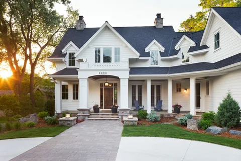 A large white two-story house with a dark roof, front porch with chairs, and a paved driveway surrounded by trees and lawn.