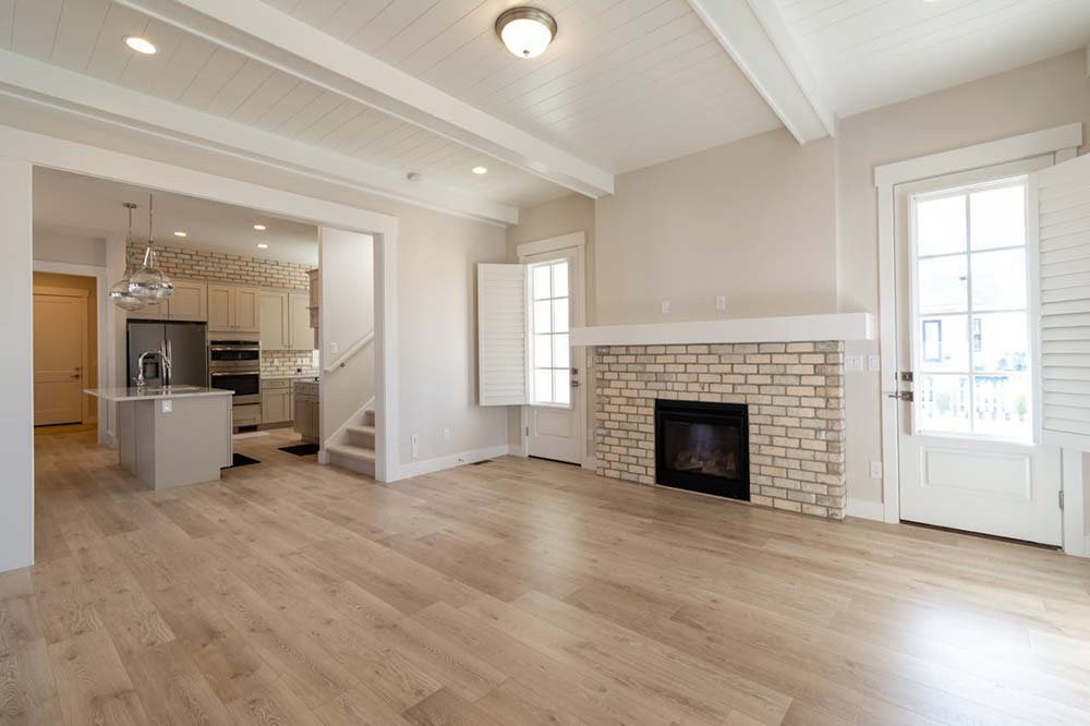 he living room of a new custom home featuring a brick fireplace, wood floors, and a view into the kitchen.