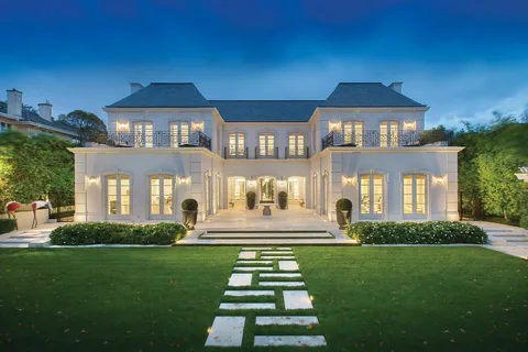 A large, modern luxury mansion with symmetrical architecture, illuminated exterior lights, and a manicured front lawn with a stone walkway leading to the entrance at dusk.