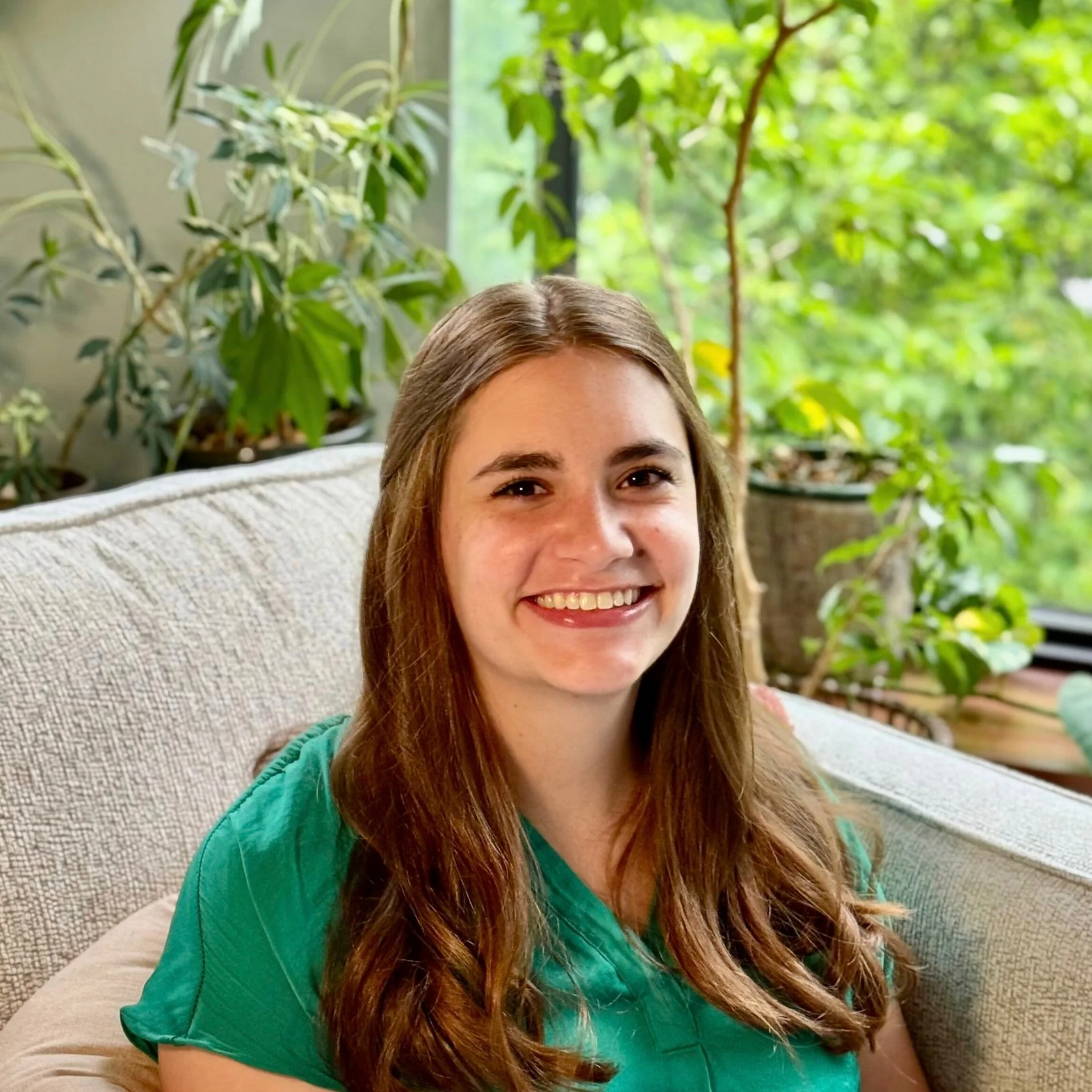 A young woman with long brown hair, smiling, sitting on a beige couch in a room with large windows and green plants in the background.