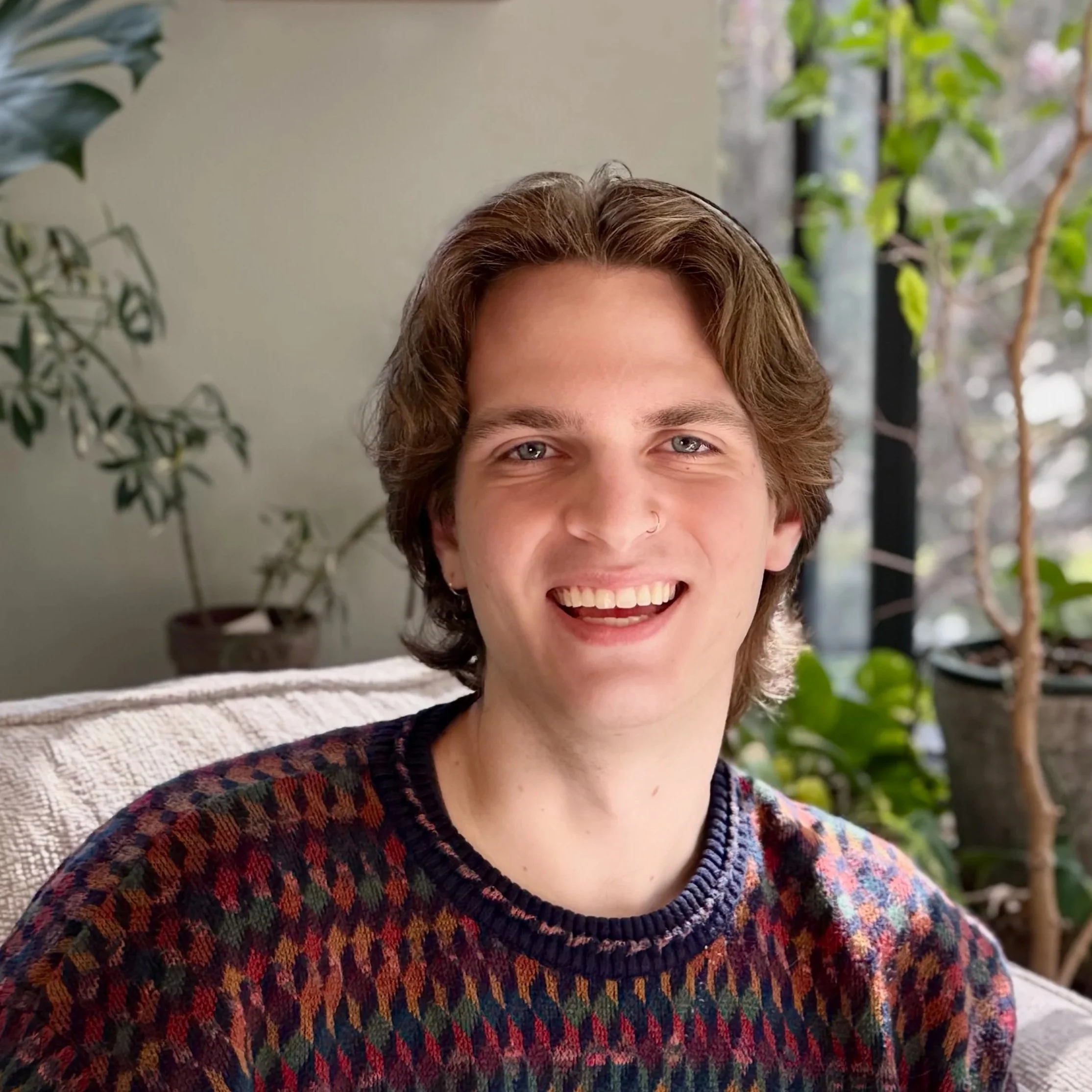 A young man with medium-length brown hair, blue eyes, and a wide smile, sitting on a beige couch with green plants and a window in the background.