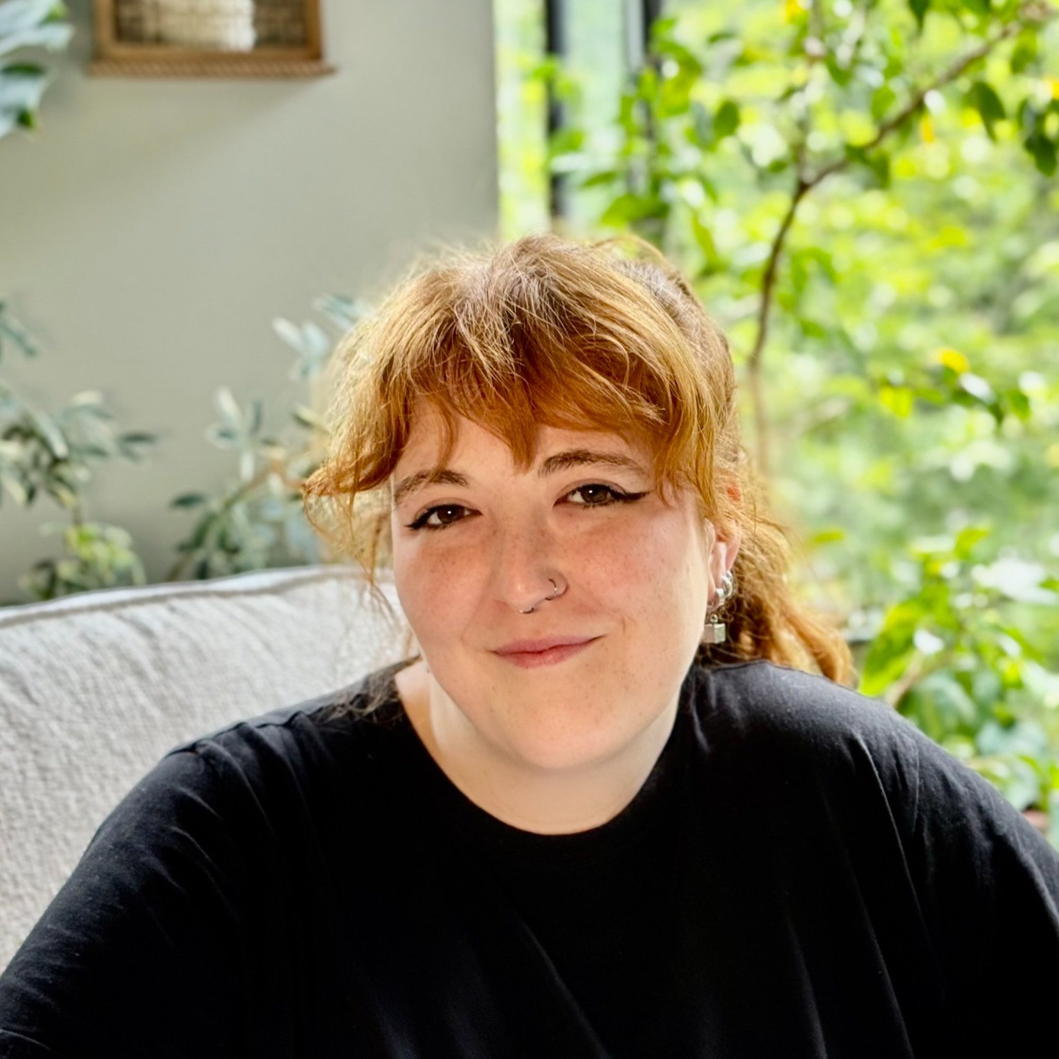 A woman with red hair, wearing a black shirt, sitting indoors near a window with green plants outside.