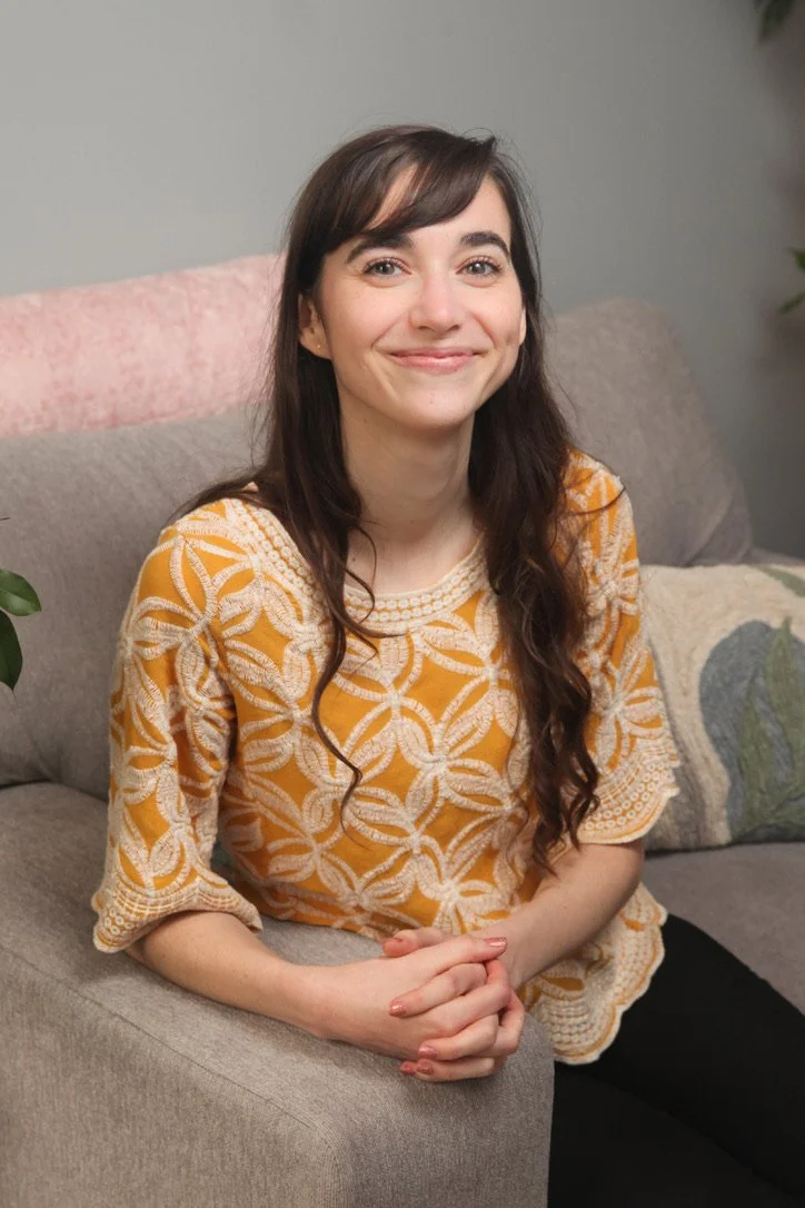 A therapist with brown hair and a yellow shirt sits on a grey chair and smiles at the camera.