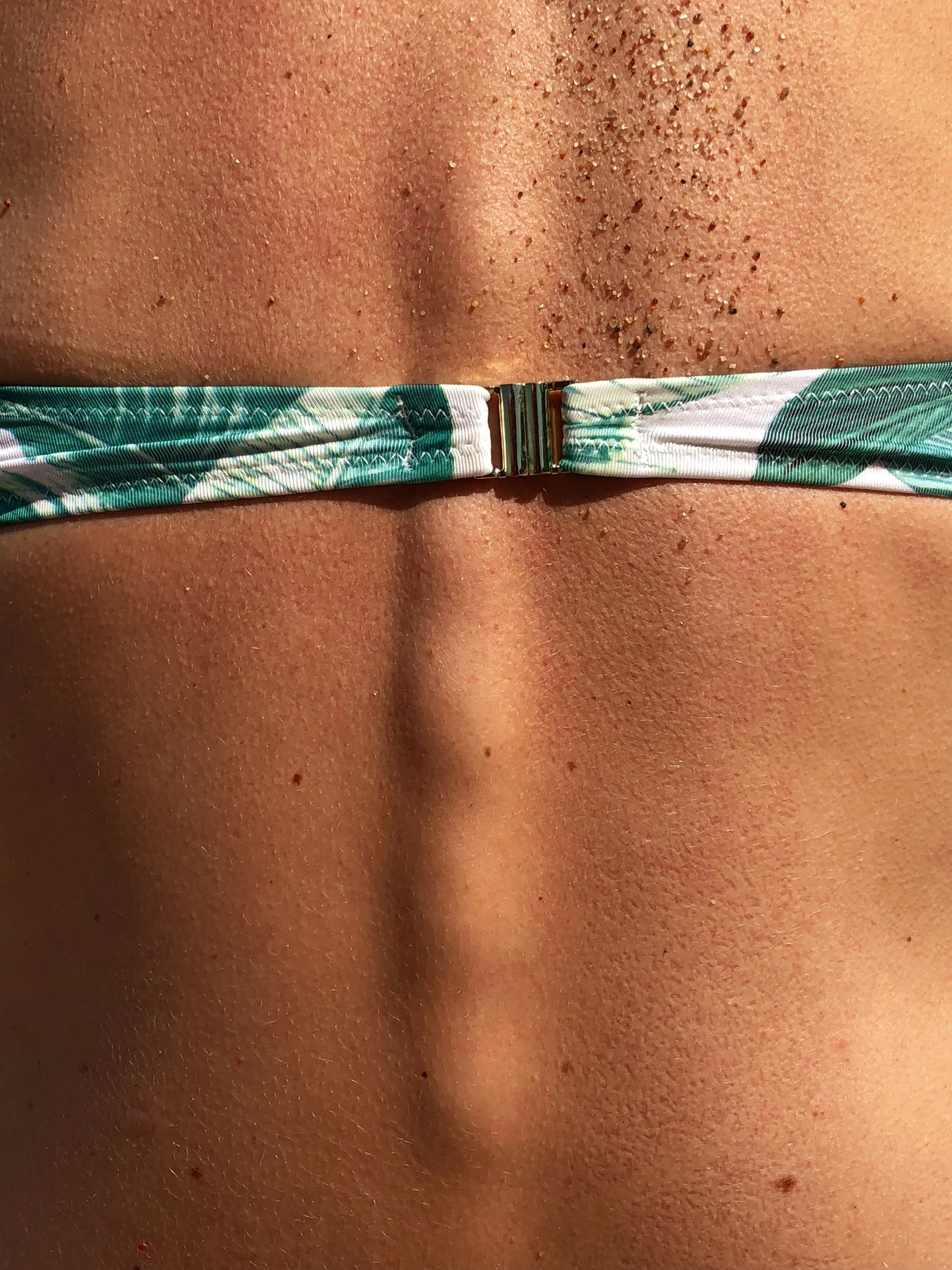 Cerca de la parte inferior de la imagen se observa tela de bikini de color verde con tonos blancos y un broche metálico en el centro, sobre piel humana.