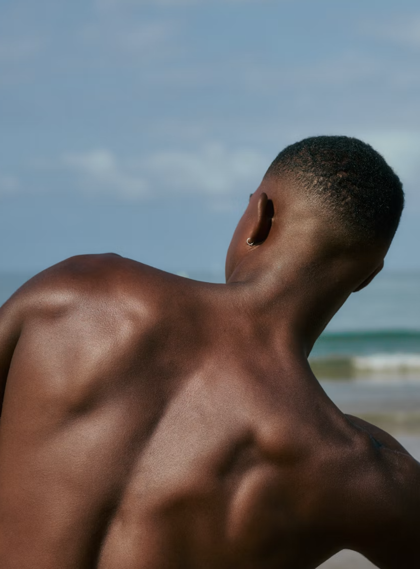 Persona con piel oscura en la playa, vista desde atrás, mostrando la espalda y el cuello, con el mar y el cielo en el fondo.