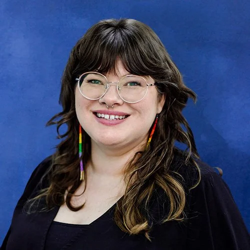 A woman with long brown hair, glasses, and rainbow earrings, smiling in a portrait against a blue background.