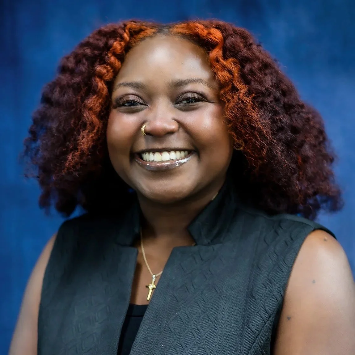 Portrait of an African American woman with long braids, wearing a black turtleneck, gold hoop earrings, and a nose ring, smiling in front of a blue background.