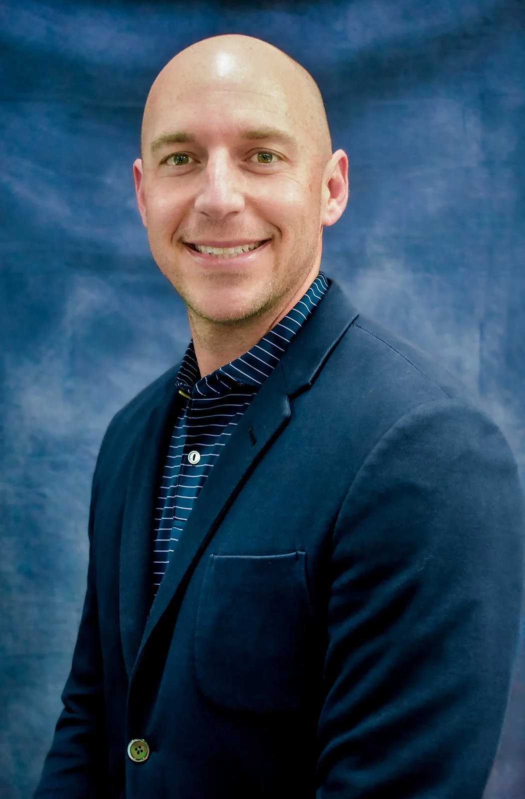 A smiling man in a dark blazer and striped shirt posing in front of a blue backdrop.