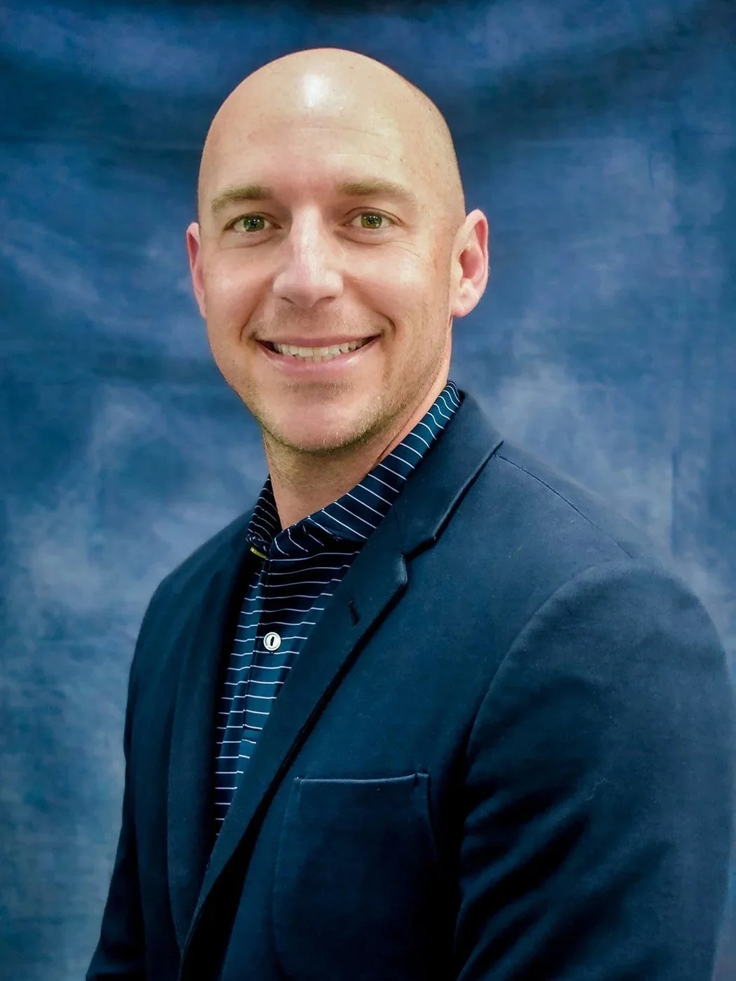 A smiling man in a dark blazer and striped shirt posing in front of a blue backdrop.