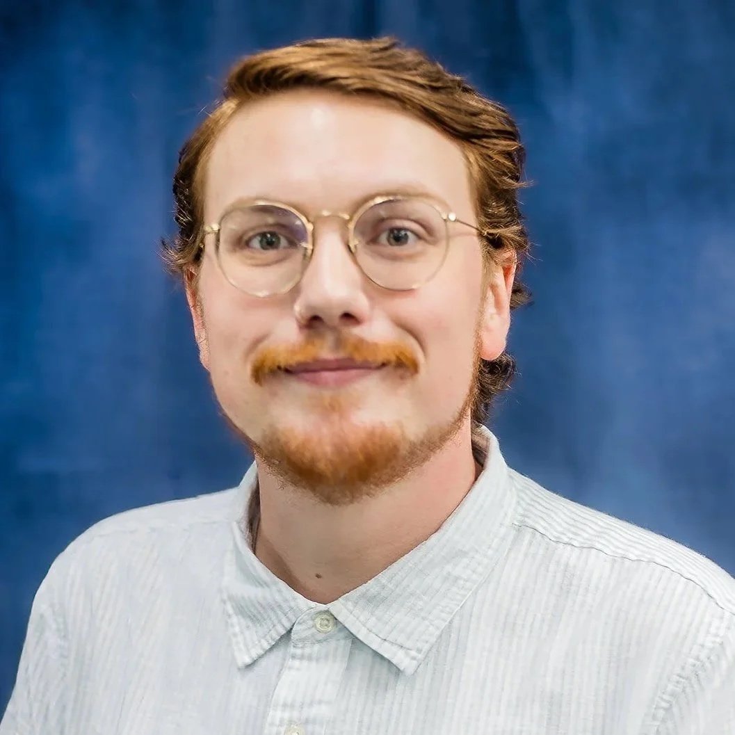 A man with glasses and a nose ring wearing a navy blazer and maroon shirt, standing in front of a blue curtain.