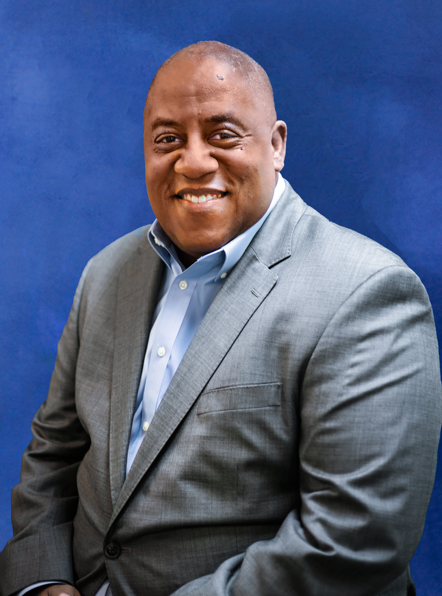 A man in a gray suit and light blue shirt smiling against a dark blue background.