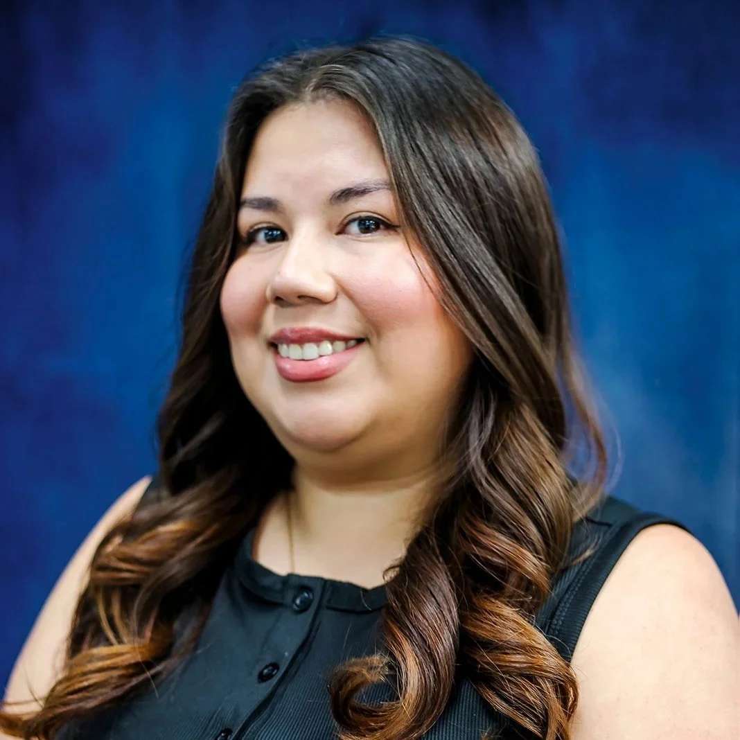 Portrait of a woman with long dark hair, smiling, wearing a brown top, black blazer, and a crystal pendant necklace, against a blue fabric backdrop.