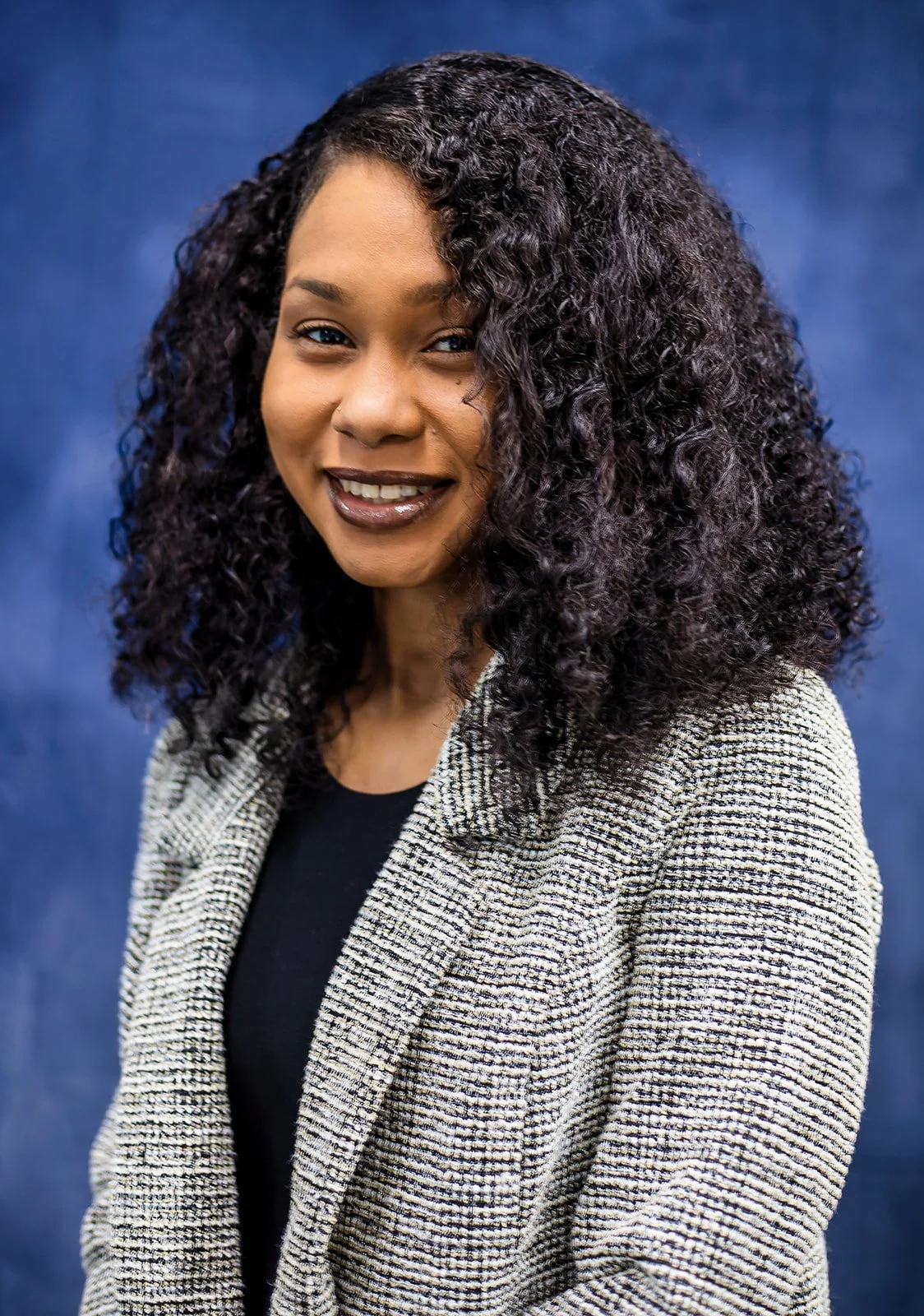 A smiling woman with curly dark hair wearing a beige checkered blazer and black top against a blue background.