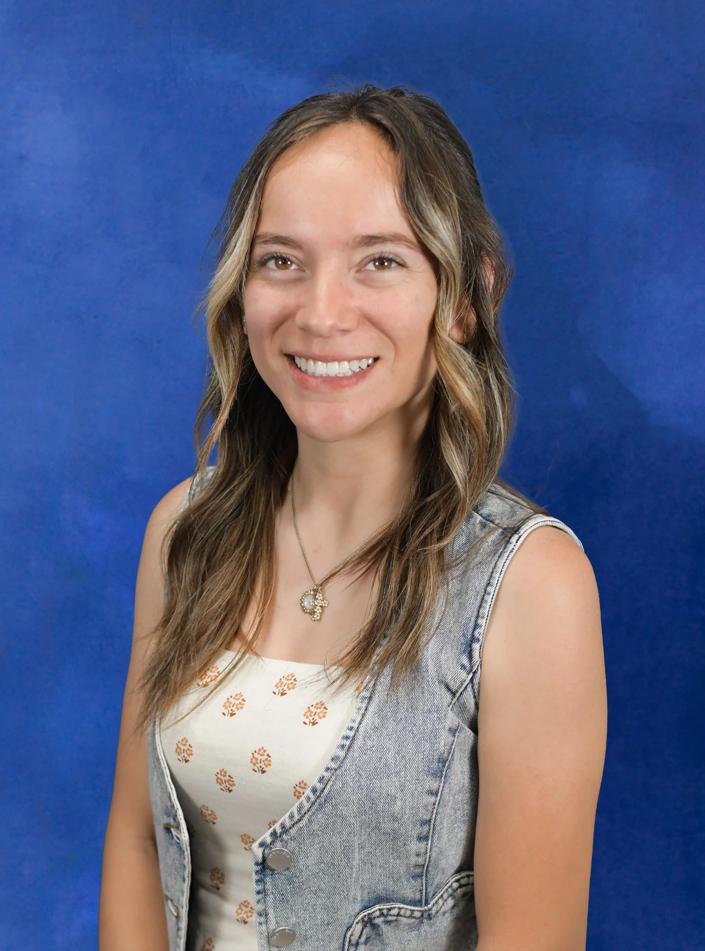 Portrait of a young woman with long brown wavy hair smiling, wearing a white long-sleeve top, gold jewelry, and standing against a blue background.