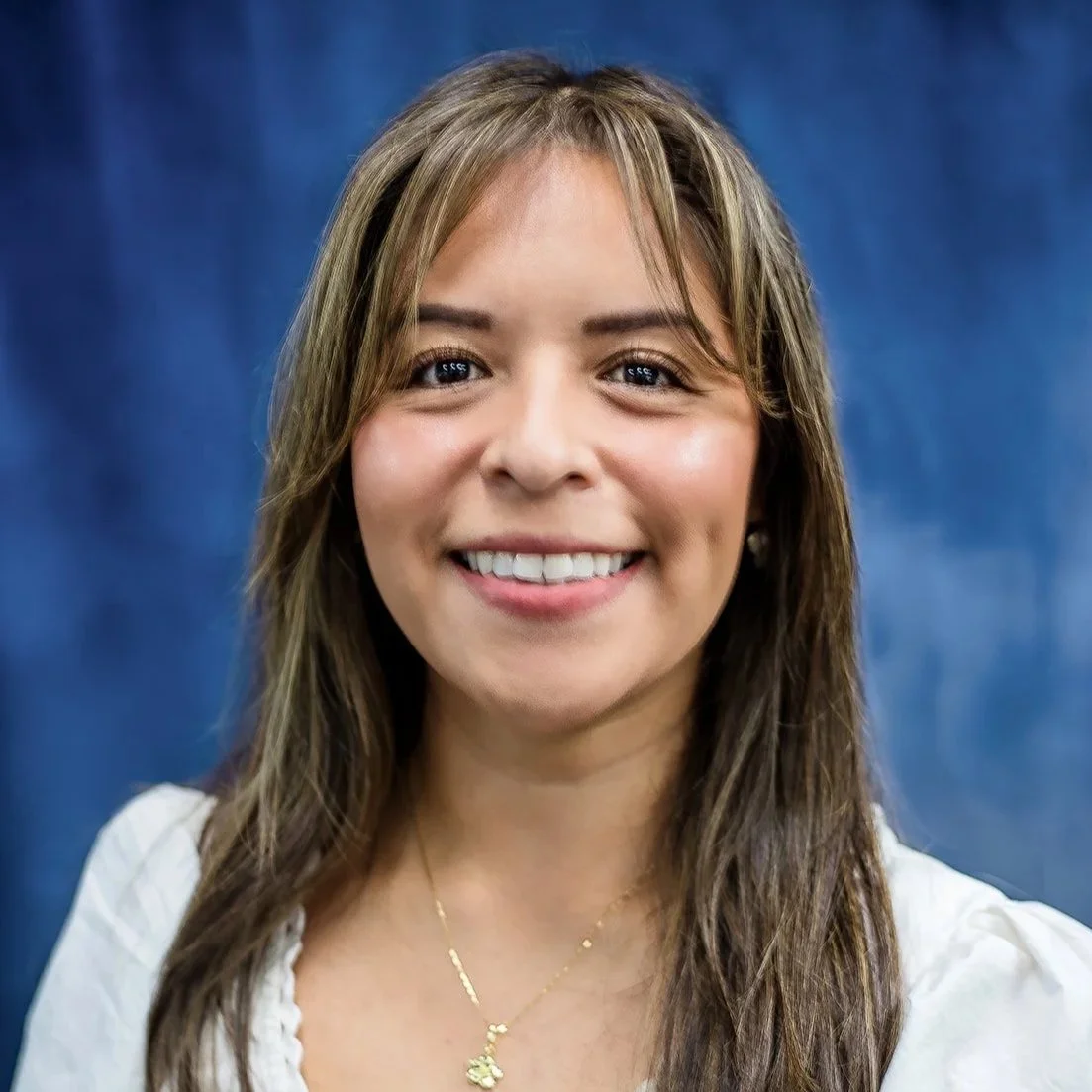Headshot of a young woman with long, dark hair smiling, wearing a brown ribbed sweater with a cutout detail at the chest, against a blue background.