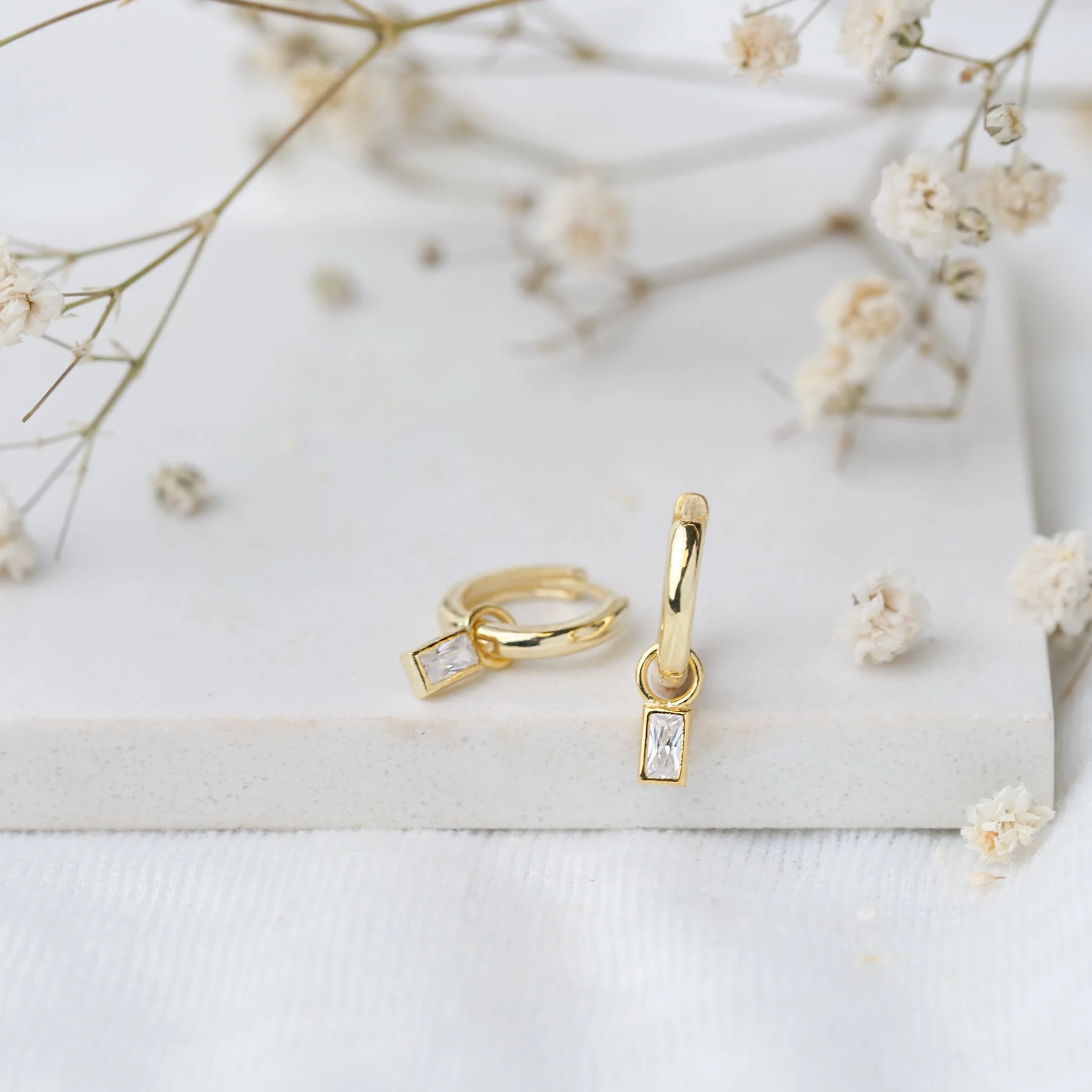 Gold hoop earrings with rectangular pendants on a white surface, surrounded by small dried flowers.