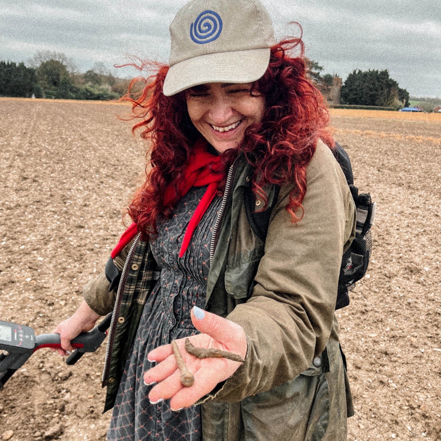 A woman with red hair wearing a baseball cap with a swirl on it and a red bandana. She is in a field, holding out her hand with some rusty nails