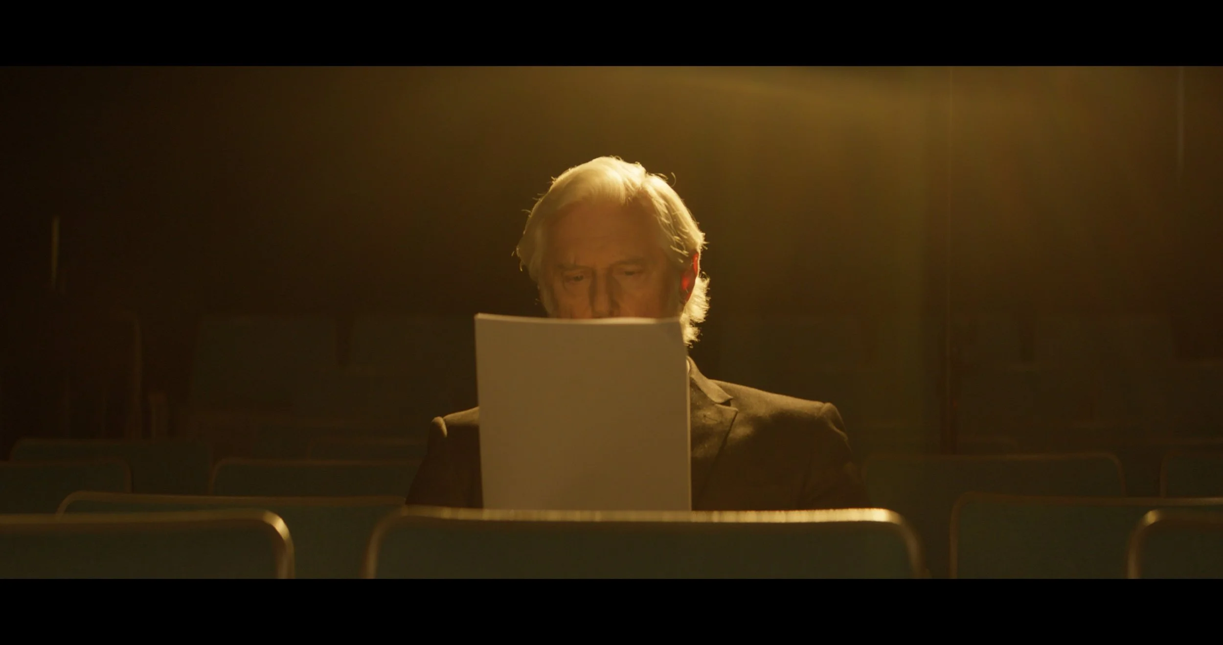 A man with gray hair is sitting alone in a dimly lit theater or auditorium, looking at a sheet of paper illuminated by a warm, yellowish light.