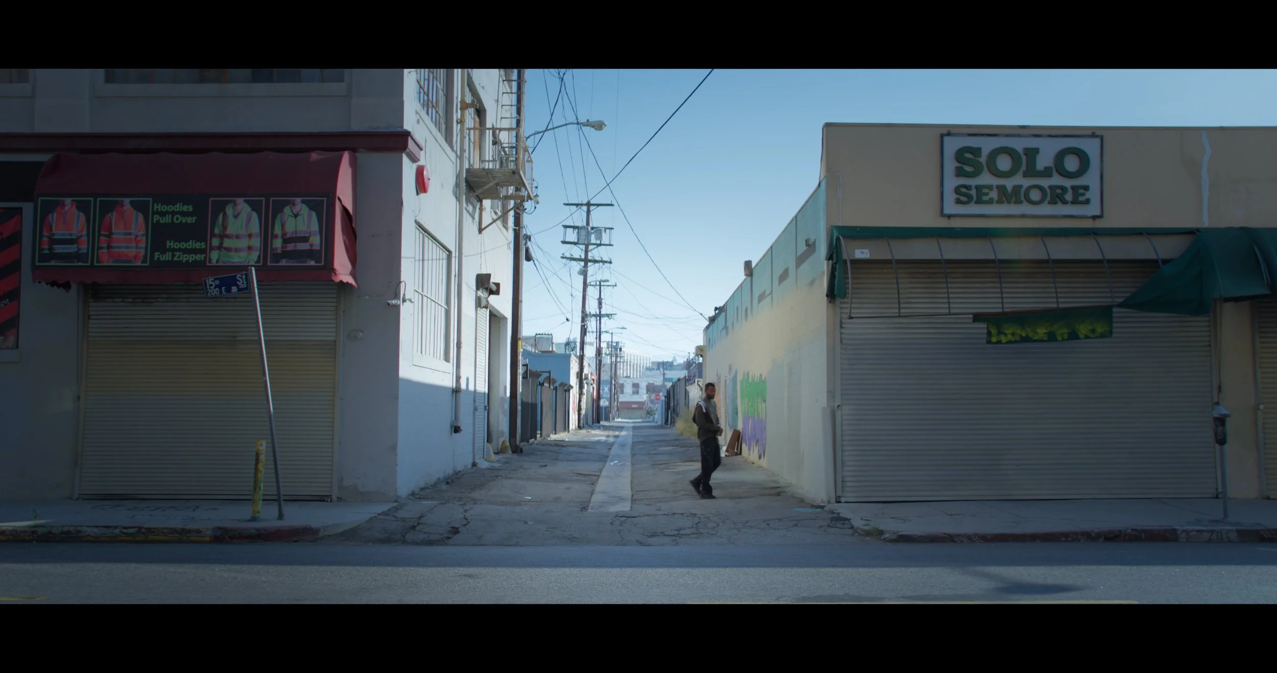An empty city street viewed from across the road, with a man standing in the alleyway between closed storefronts, some graffiti on the walls, and utility poles with wires overhead.