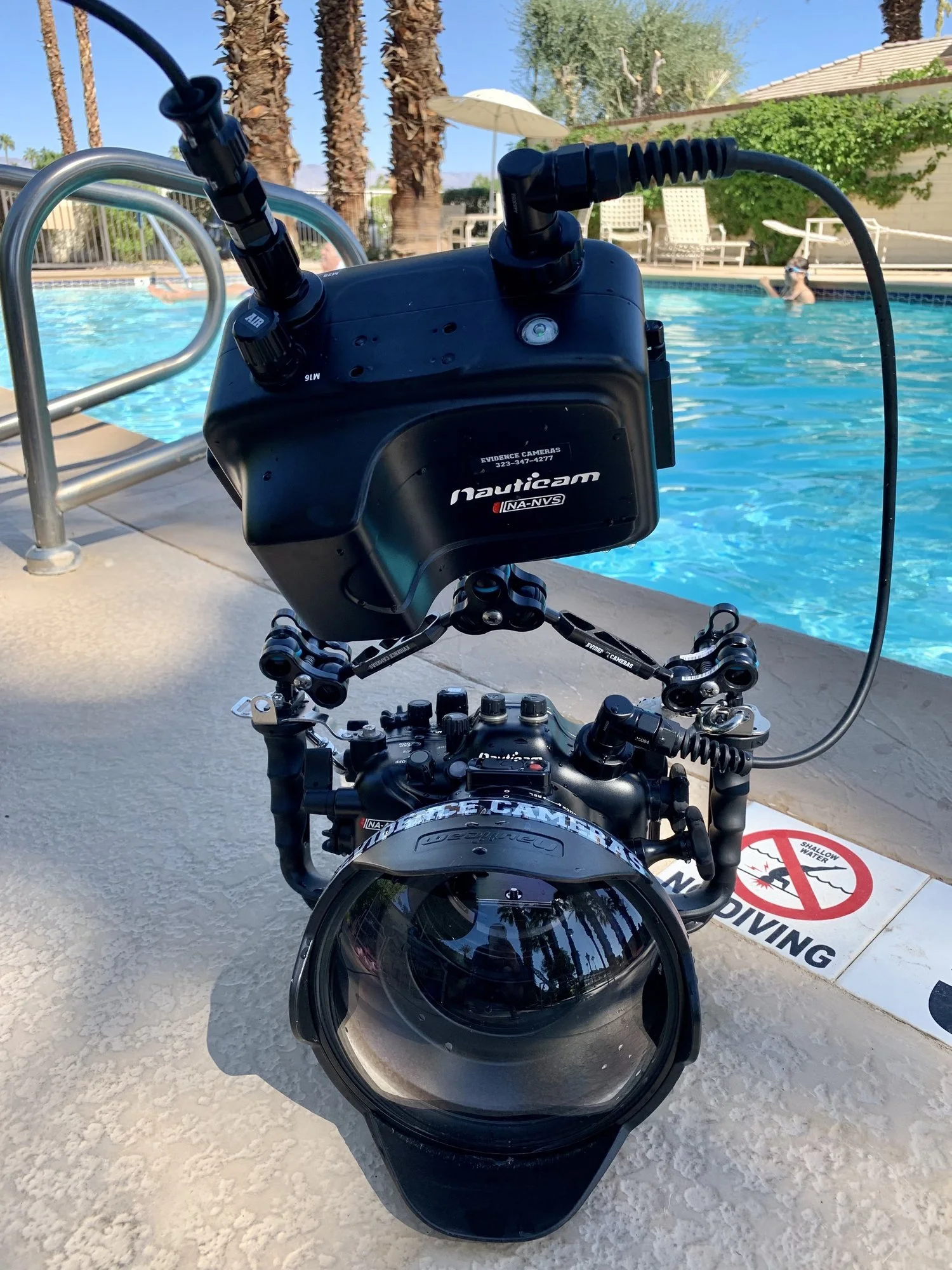A diver's helmet attached to a camera rig is positioned near the swimming pool edge, with people swimming in the background and a no diving sign on the ground.
