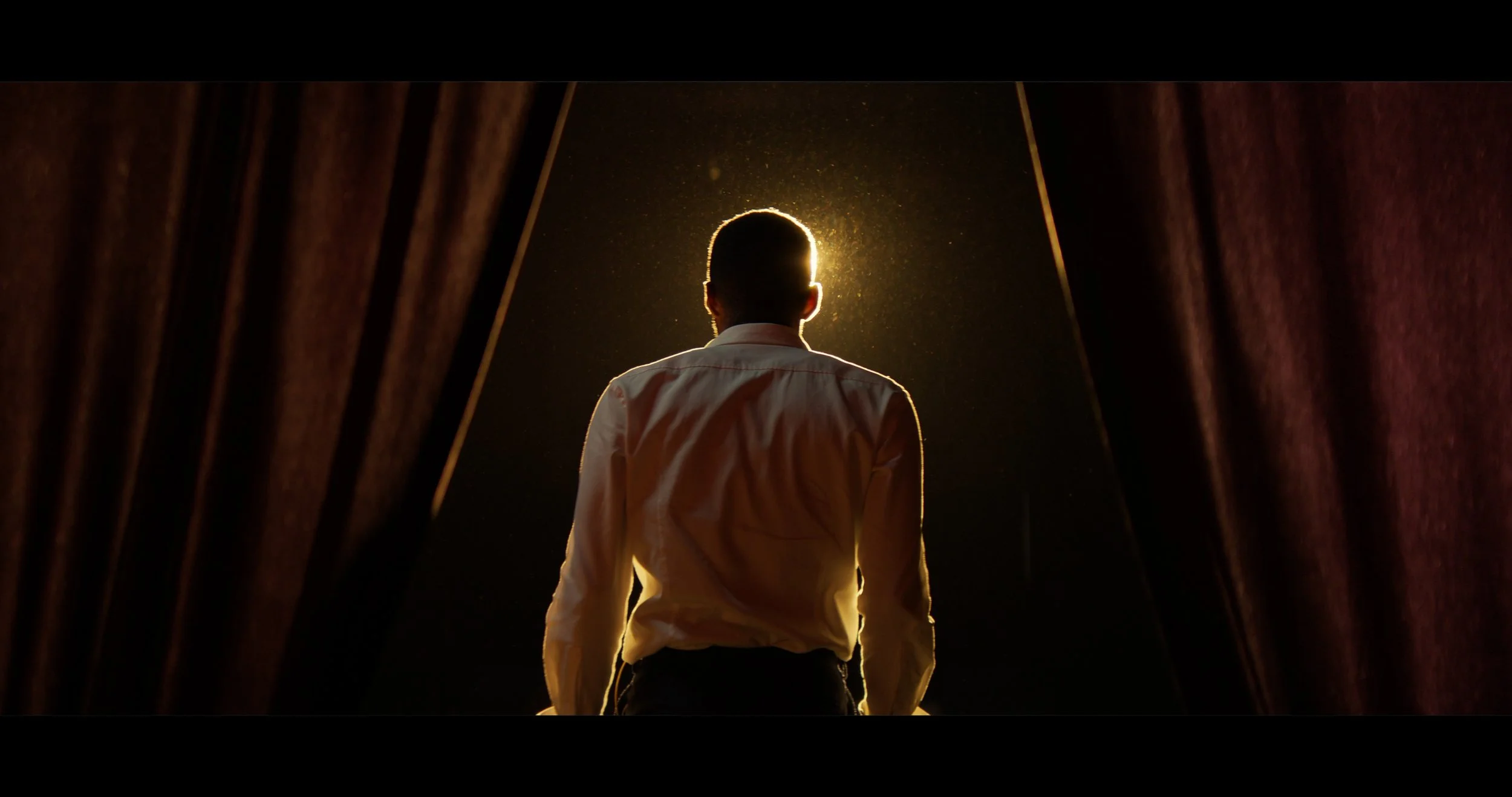 A person with short hair wearing a white shirt standing at the edge of a stage or opening, looking towards a bright light or moon in the dark sky, with curtains on either side.