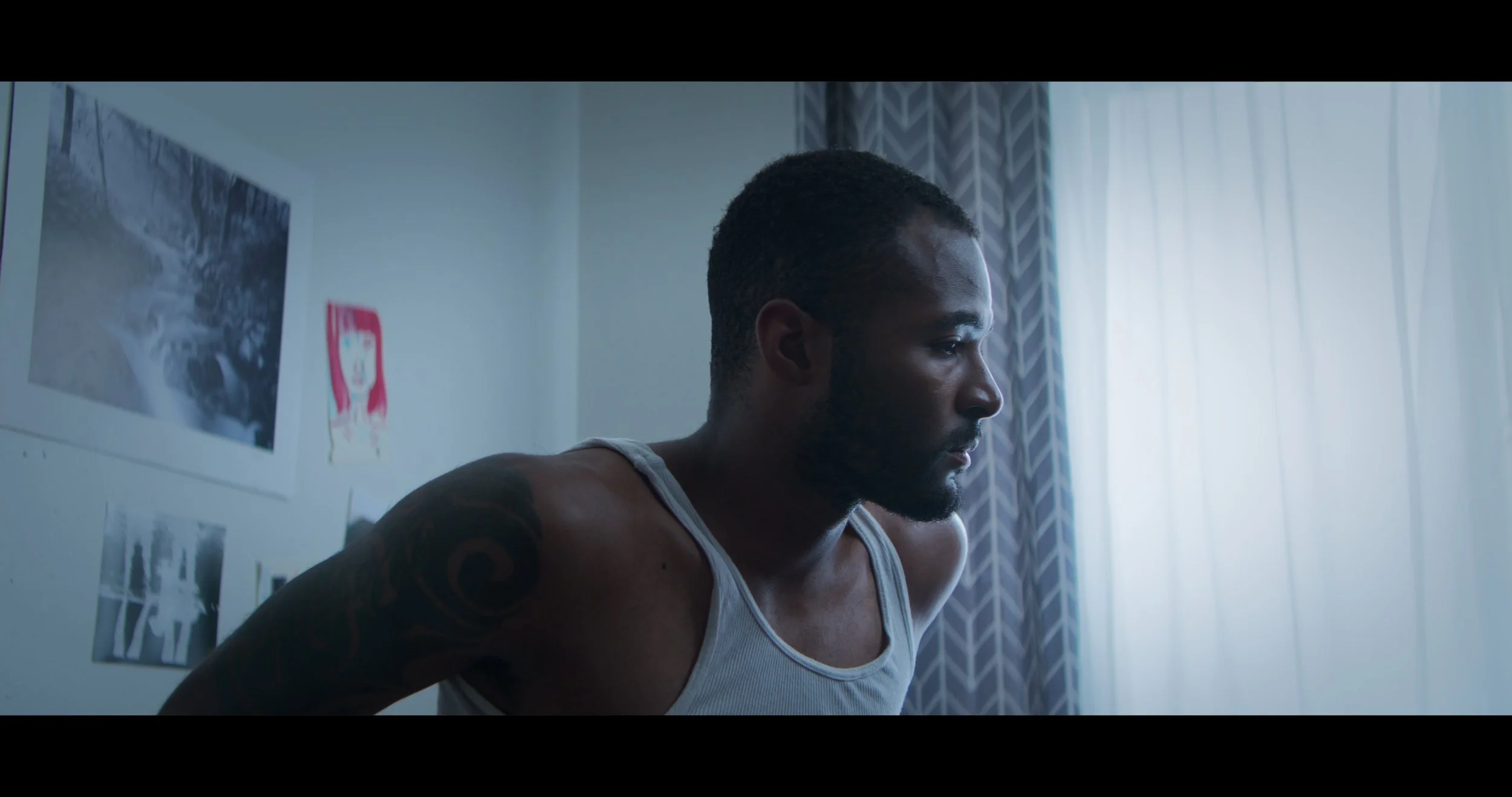 A man with tattoos in a gray tank top leaning forward in a room with blue curtains and wall art, looking intently to the right.