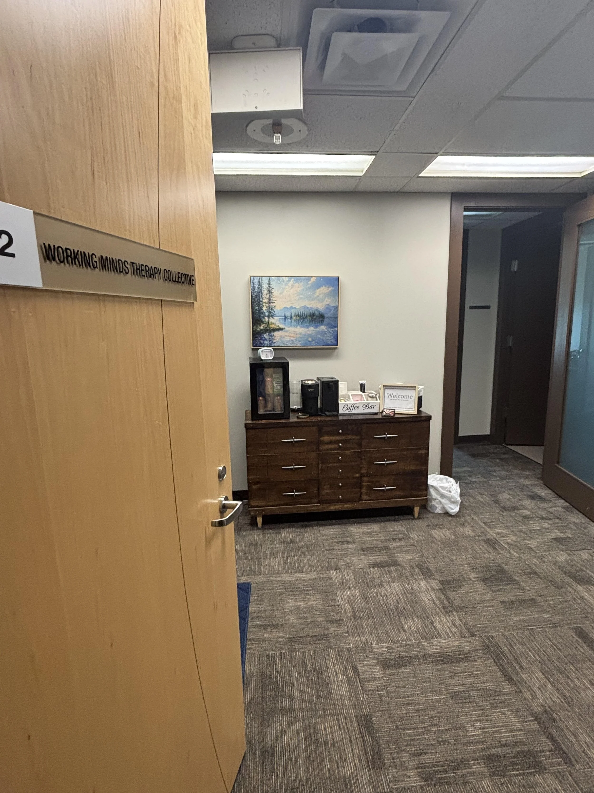 Interior of an office workspace with a wooden door on the left labeled 'WORKING MINDS THERAPY COLLECTIVE.' Inside, there is a small wooden cabinet with various items on top, a framed picture of a lake scene on the wall, and a coffee station. The room has white walls, a bookshelf, and a doorway leading to another room.