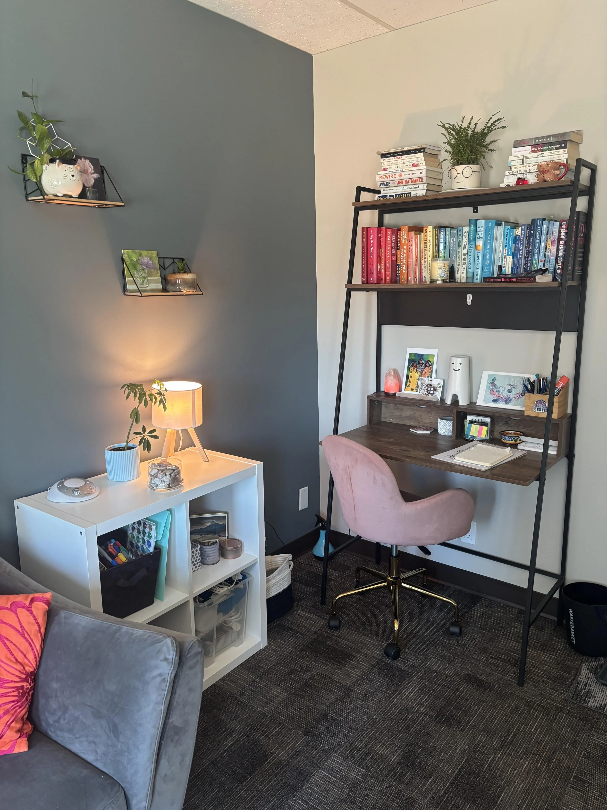 A corner of a home office with a gray couch, pink pillow, white shelf with office supplies, a black desk with books, a pink chair, a bookshelf with colorful books, and decorative items, illuminated by a small table lamp and wall lighting.