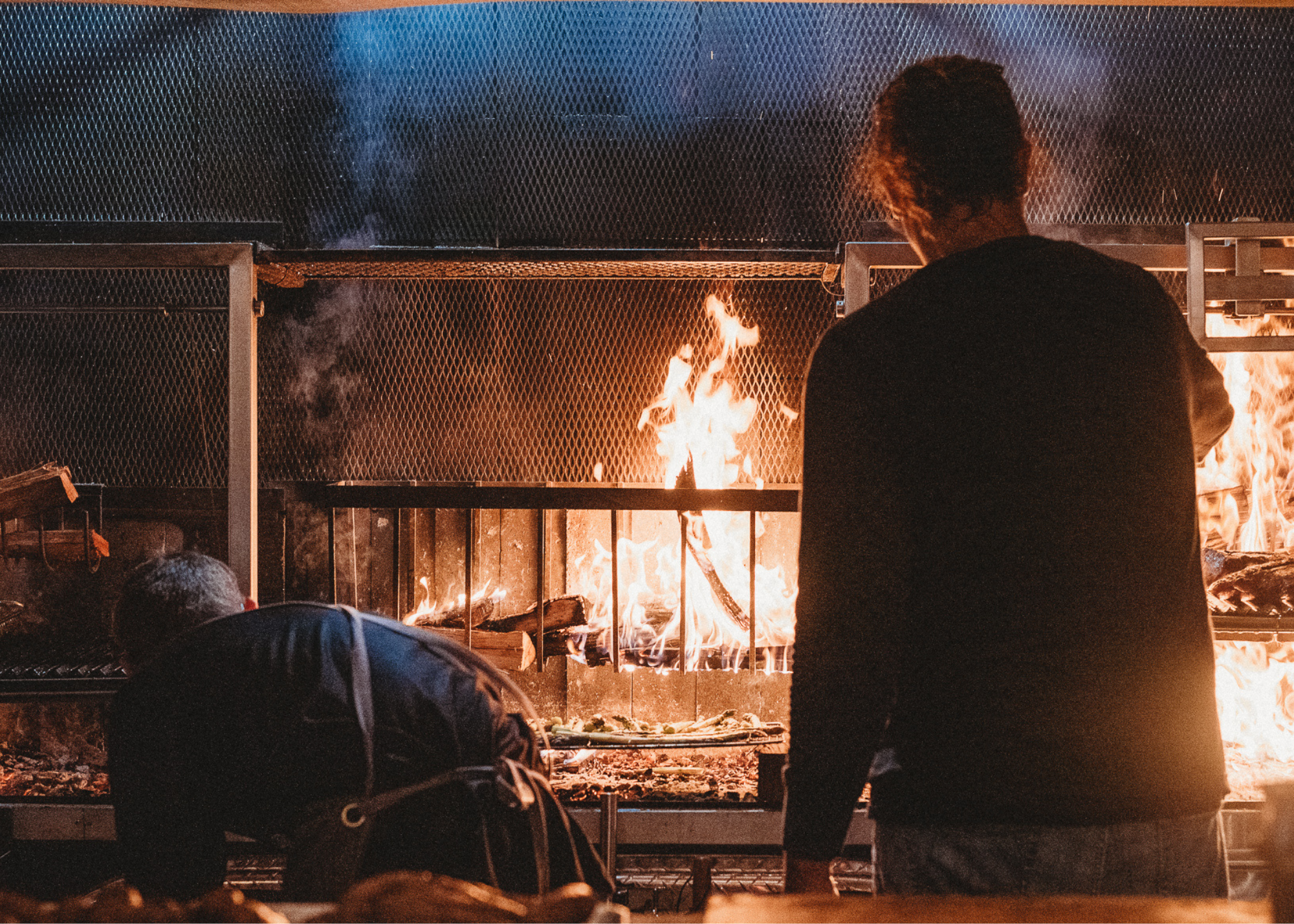 Two chefs cooking on a backlit live fire parrilla grill