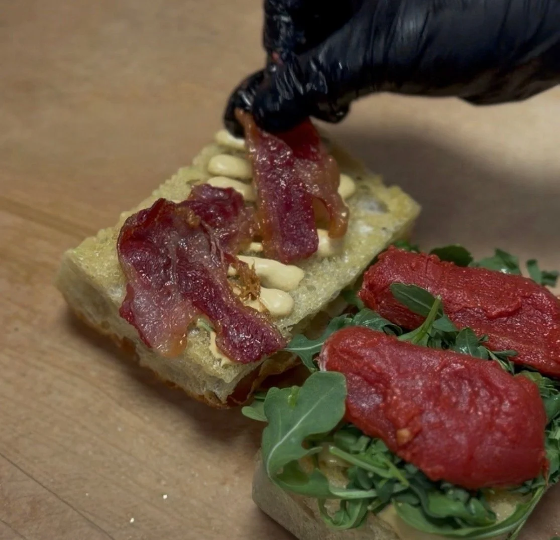Housemade beef bacon being placed onto a freshly prepared sandwich during assembly.