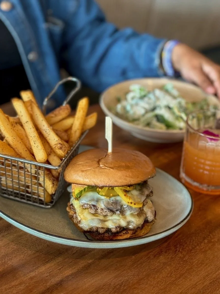 Smash Burger plated with a basket of fries and a Caesar salad and cocktail in the background