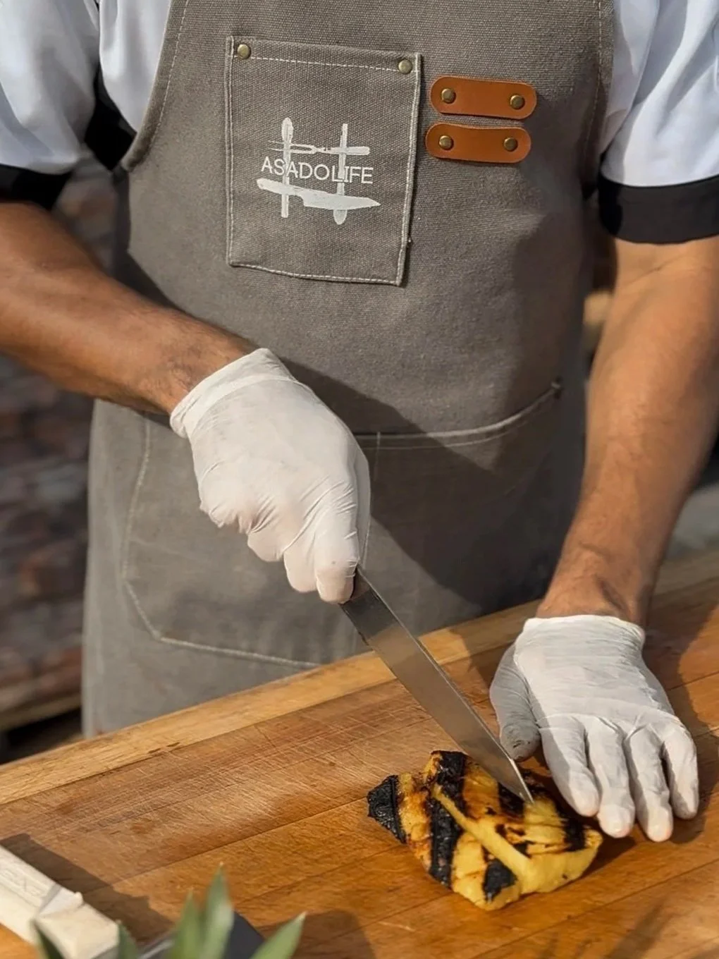 Chef in a branded Asado Life apron cutting a pineapple with grill marks on a cutting board