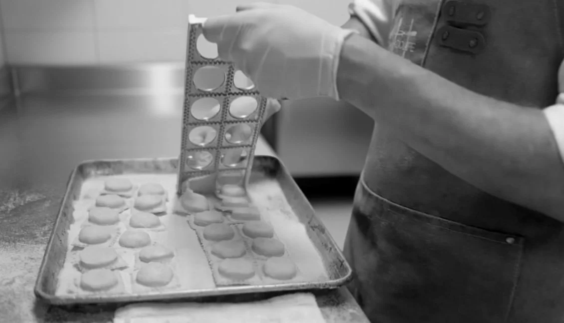 A gloved person in an apron holds a metal cutter tray over a baking sheet, releasing small, round pieces of dough arranged neatly on a floured surface.