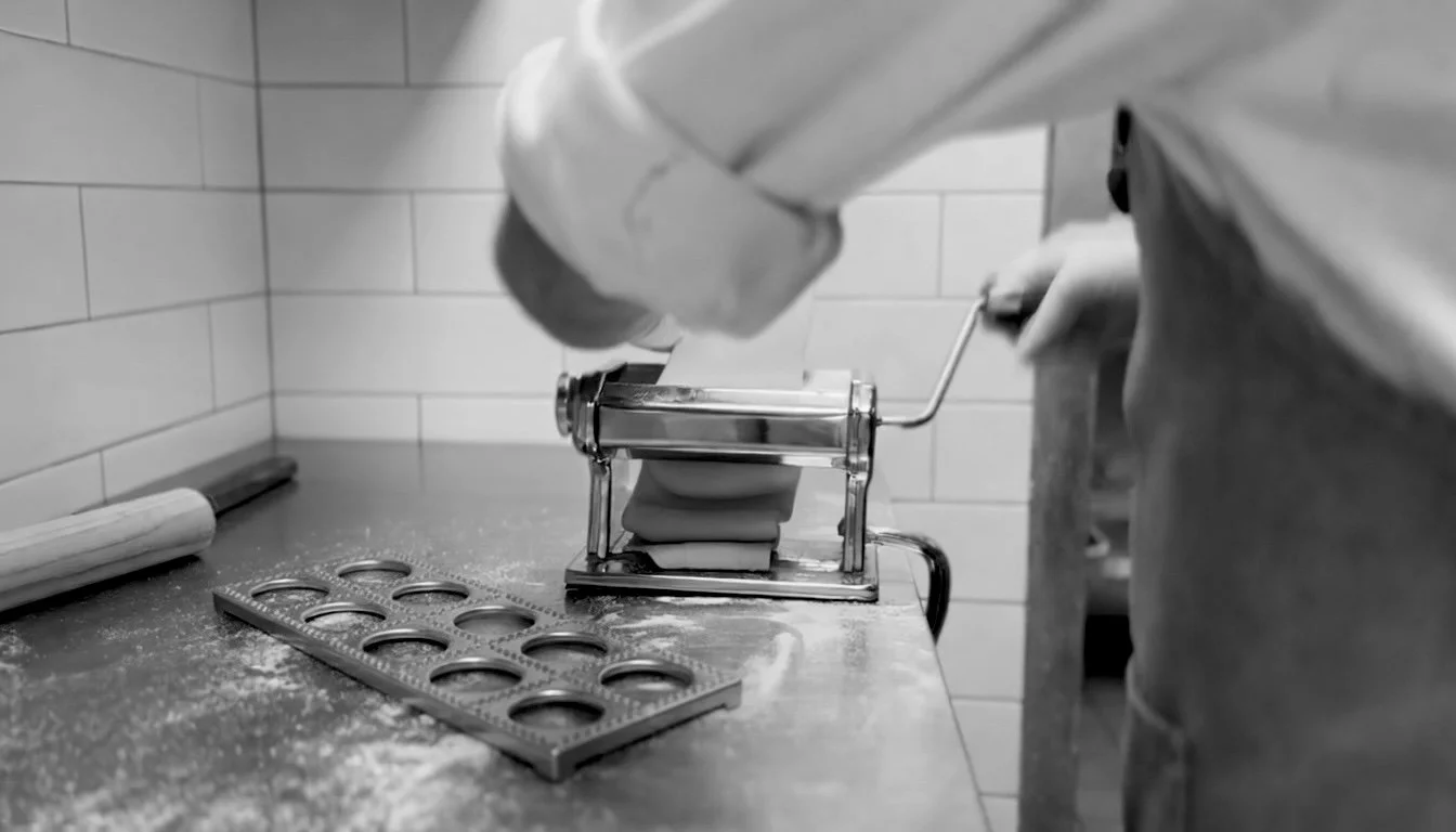 A person wearing gloves uses a hand-cranked pasta machine on a floured countertop, rolling out dough in a tiled kitchen workspace. A metal mold and rolling pin sit nearby.