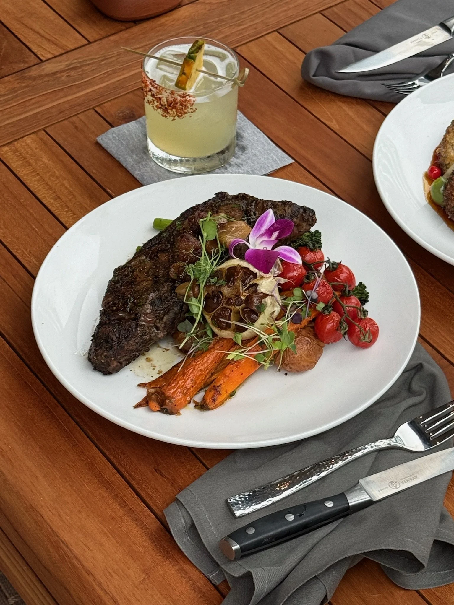 A white plate with cooked meat garnished with edible flowers, microgreens, cherry tomatoes, and roasted carrots on a wooden table.