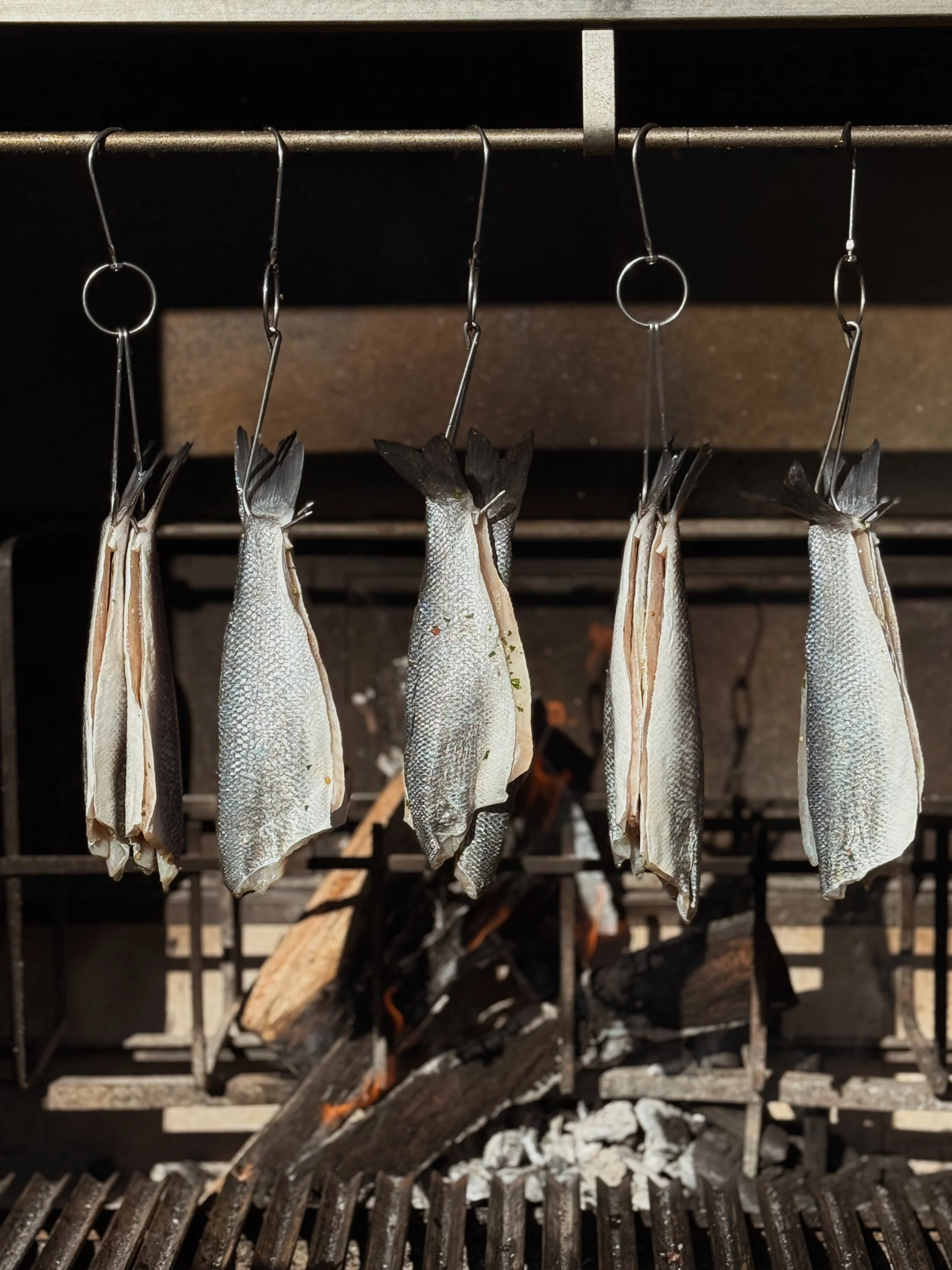 Bronzino filets hanging by S-hooks over a live fire