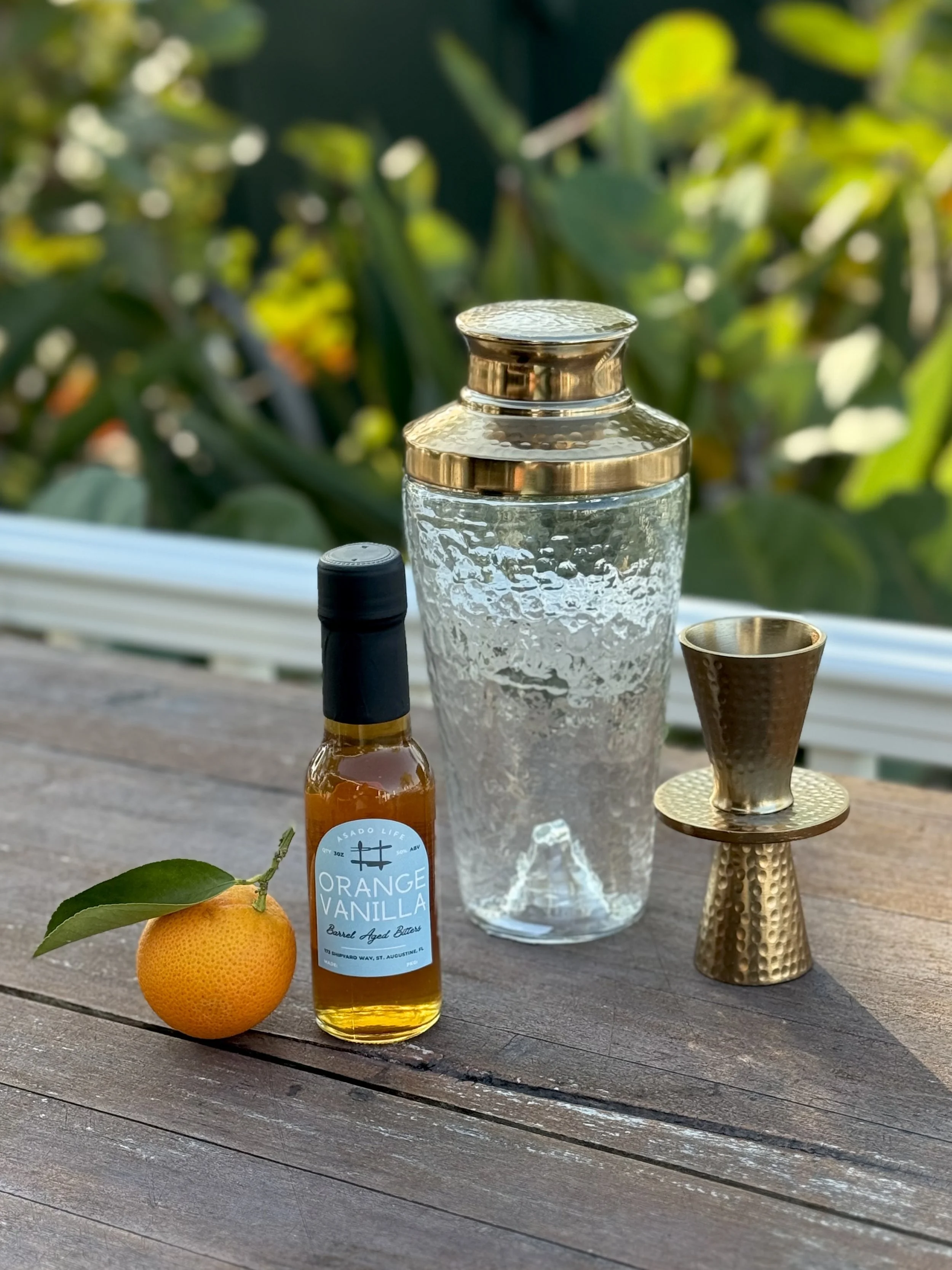 Orange Vanilla Bitters in bottle next to a mini orange, cocktail shaker, and jigger