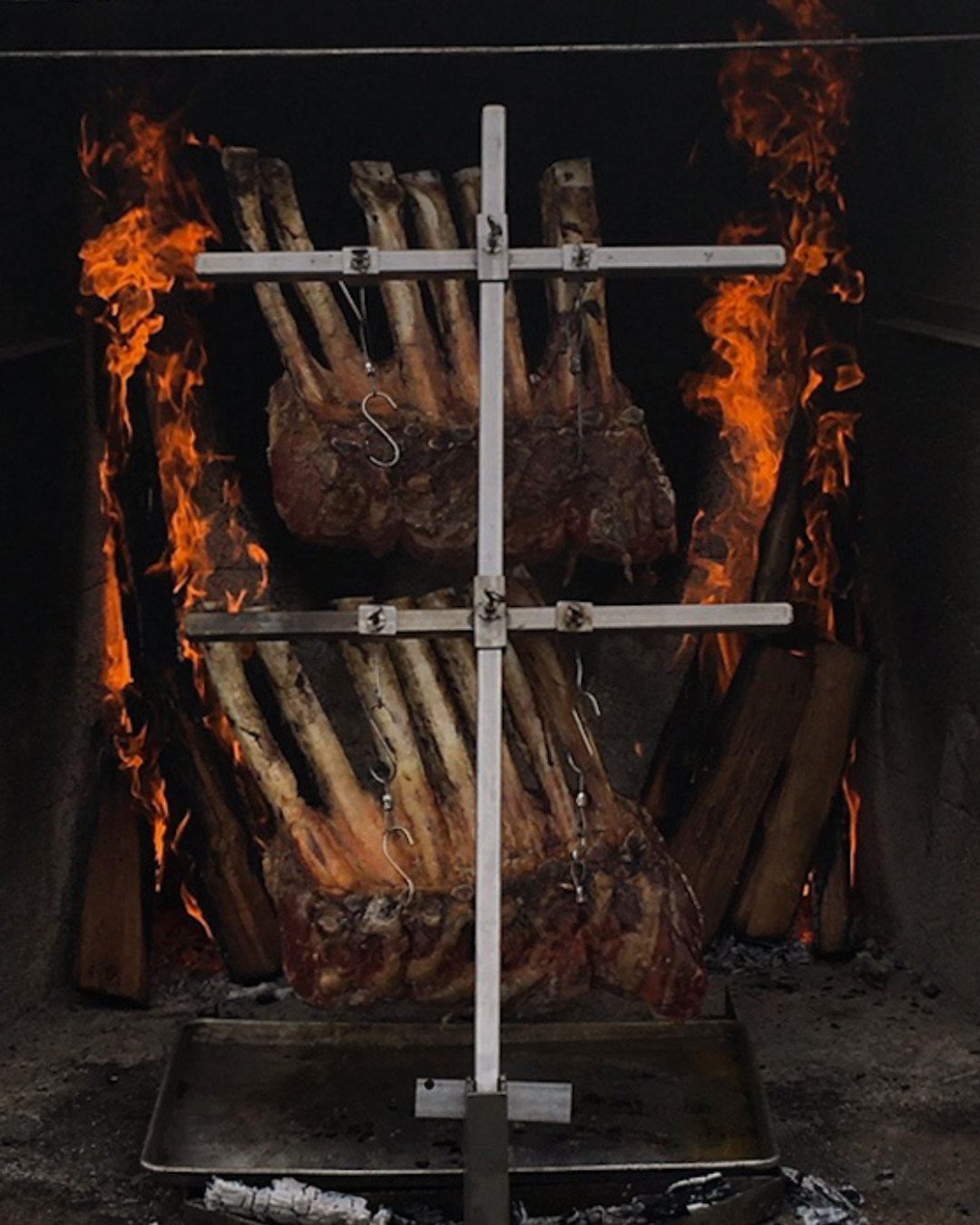 A large piece of meat being cooked over an open flame on a spit with wood logs burning on the sides.