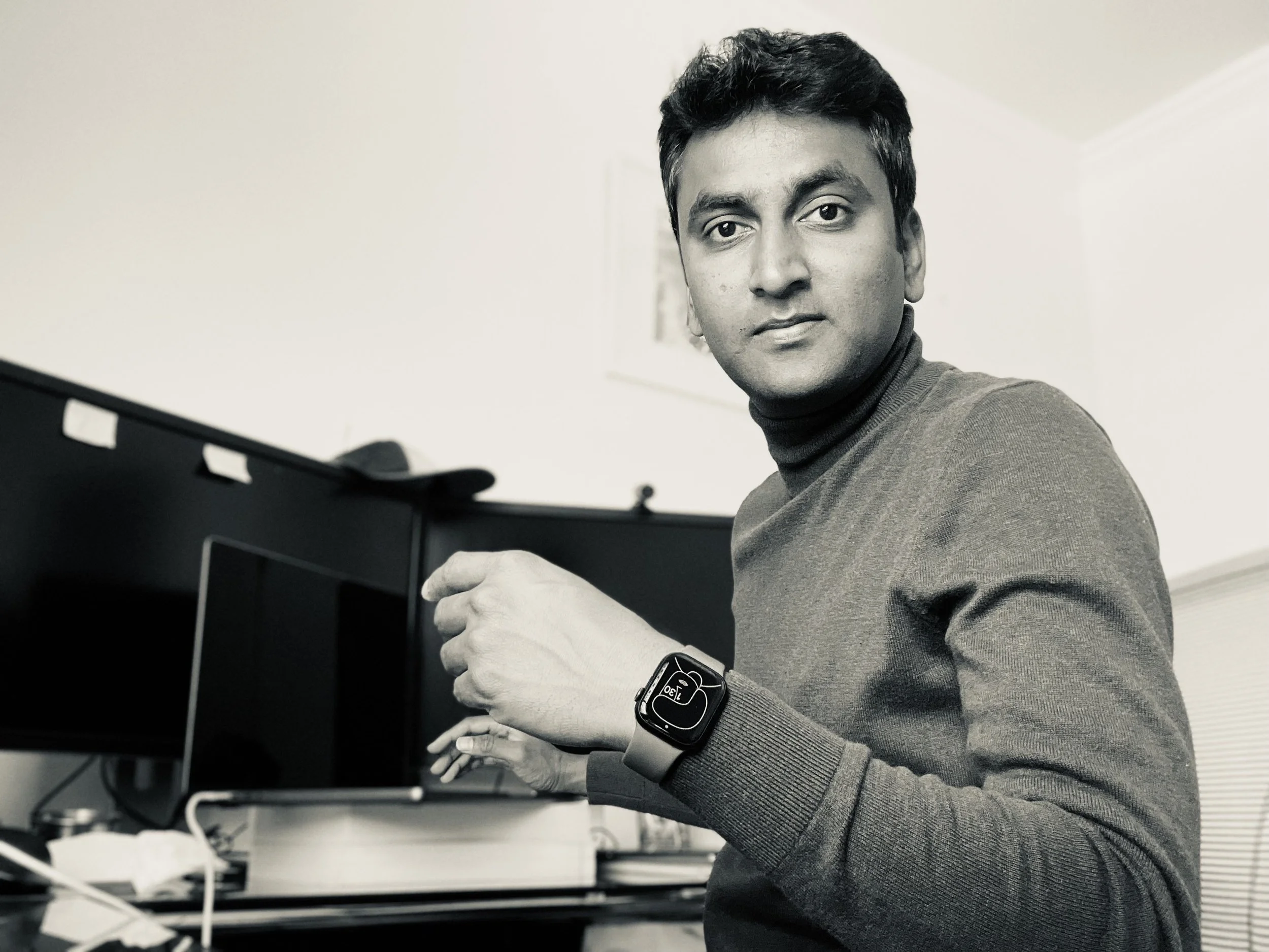 A man with short dark hair and a neutral expression is sitting at a desk, wearing a dark turtleneck sweater and a smartwatch on his left wrist. He is holding his right hand near a computer keyboard in a room with light-colored walls.