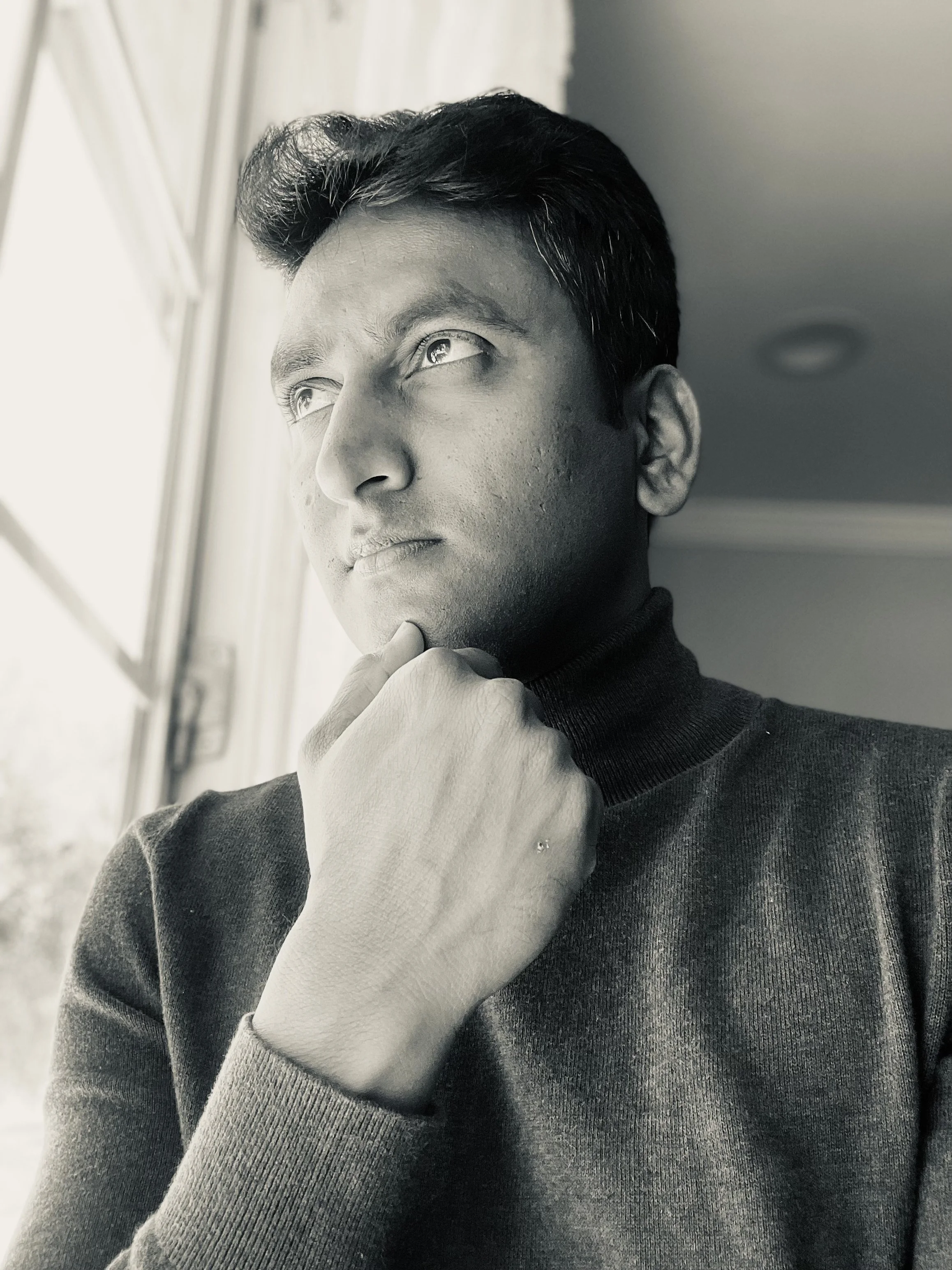 Black and white portrait of a young man looking out a window with his hand on his chin, wearing a dark turtleneck sweater.