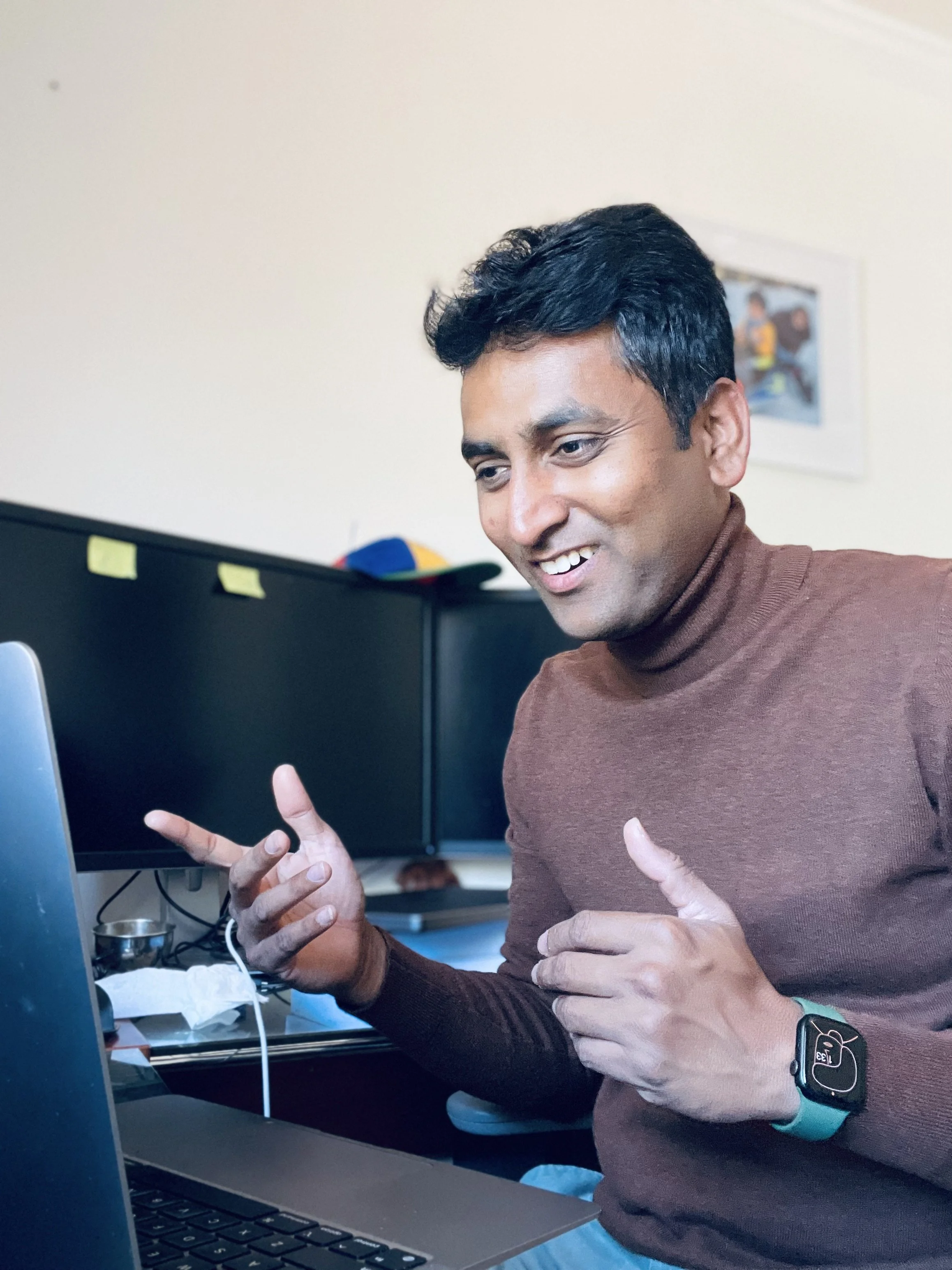 A man with dark hair wearing a brown sweater, smiling and giving a peace sign with his fingers, sitting in front of a computer.