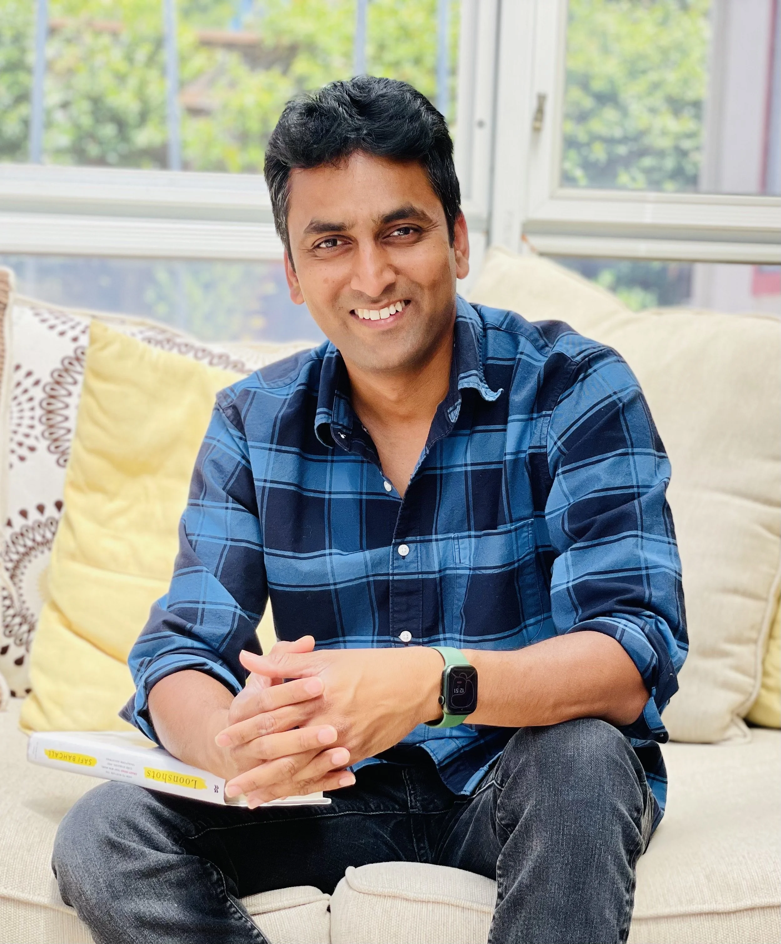 A man sitting on a beige sofa, smiling, wearing a blue plaid shirt, dark jeans, and a smartwatch. There are yellow pillows behind him and a newspaper on his lap. In the background, there are large windows with a view of greenery outside.