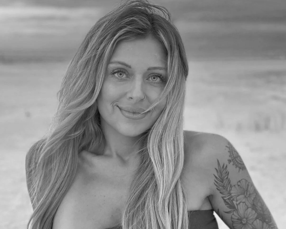 A woman with long wavy hair and tattoos on her arm, smiling at the camera on a beach in black and white.