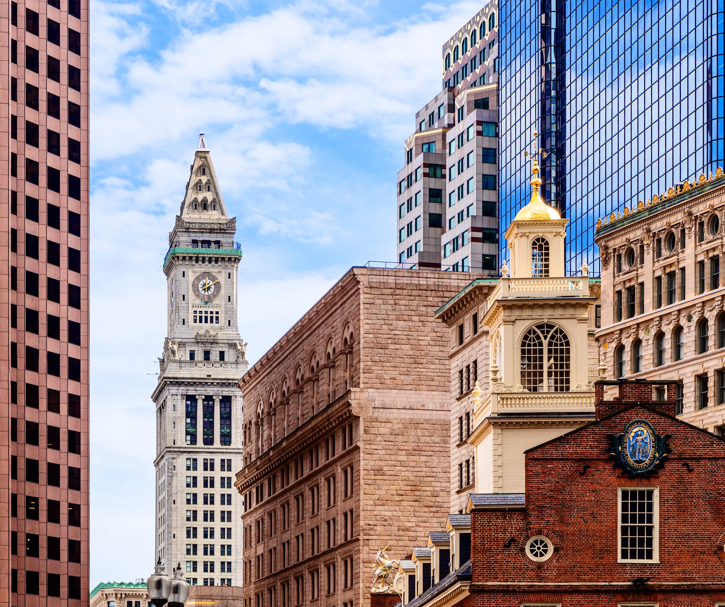 View of the cream-colored clock tower among other historic brick and modern tall glass buildings in downtown Boston.