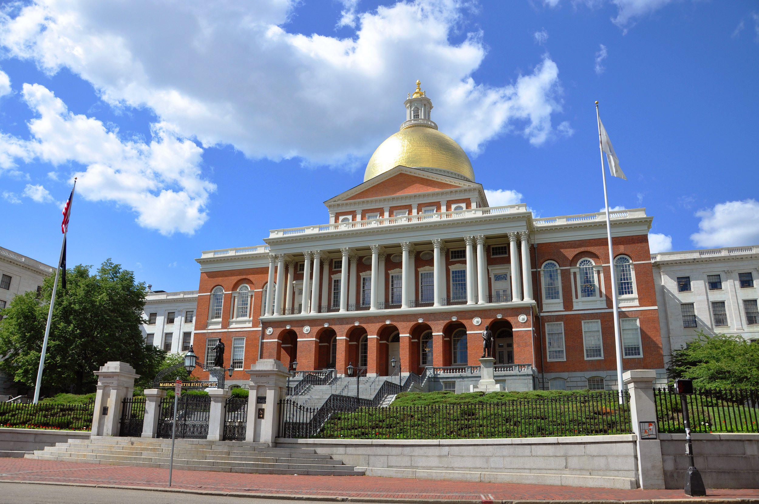 Modern photograph of the Massachusetts State House, designed by Charles Bulfinch, showing its exterior and golden dome against a blue sky with white clouds.