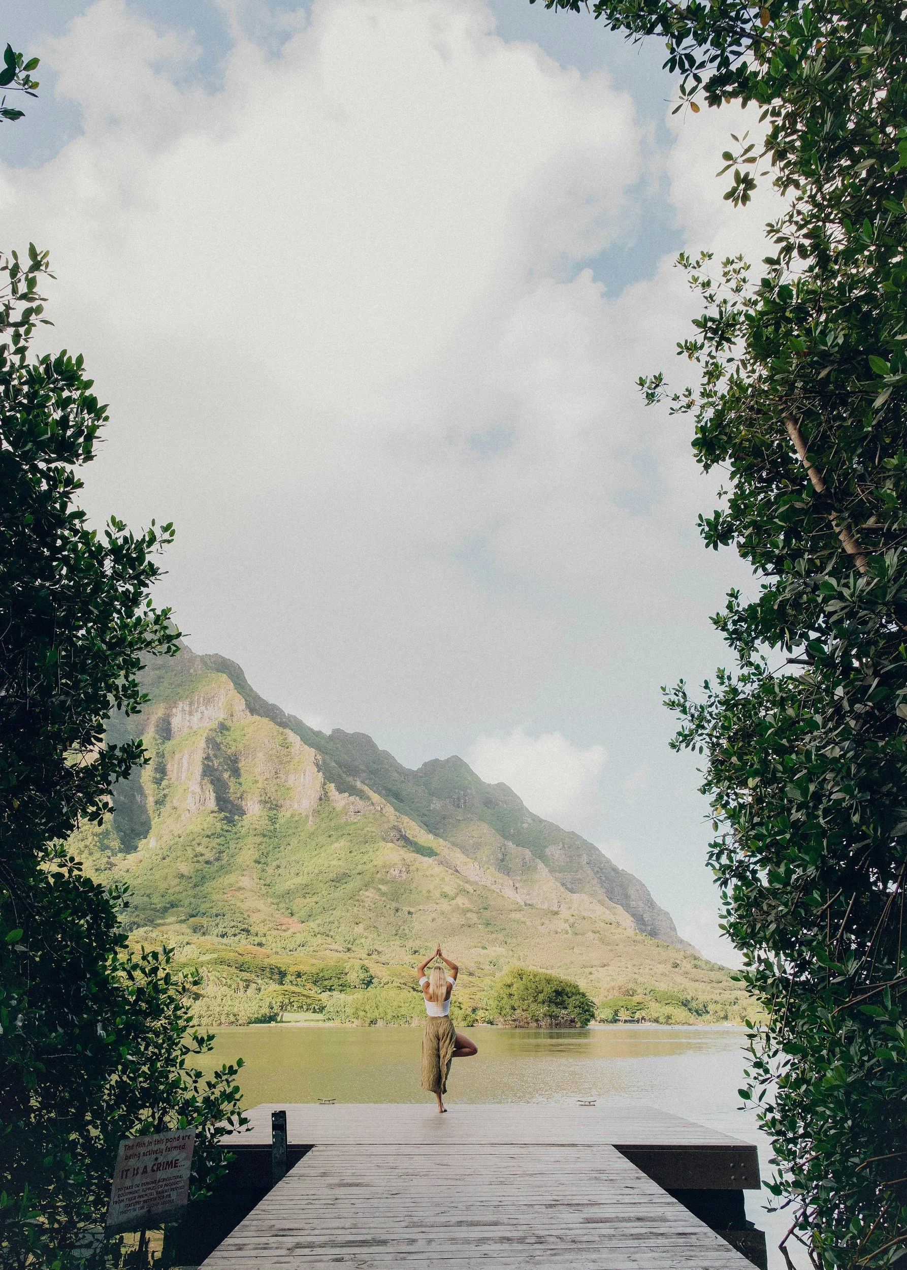 A person practicing yoga on a dock by a lake, surrounded by green trees and mountains under a cloudy sky.