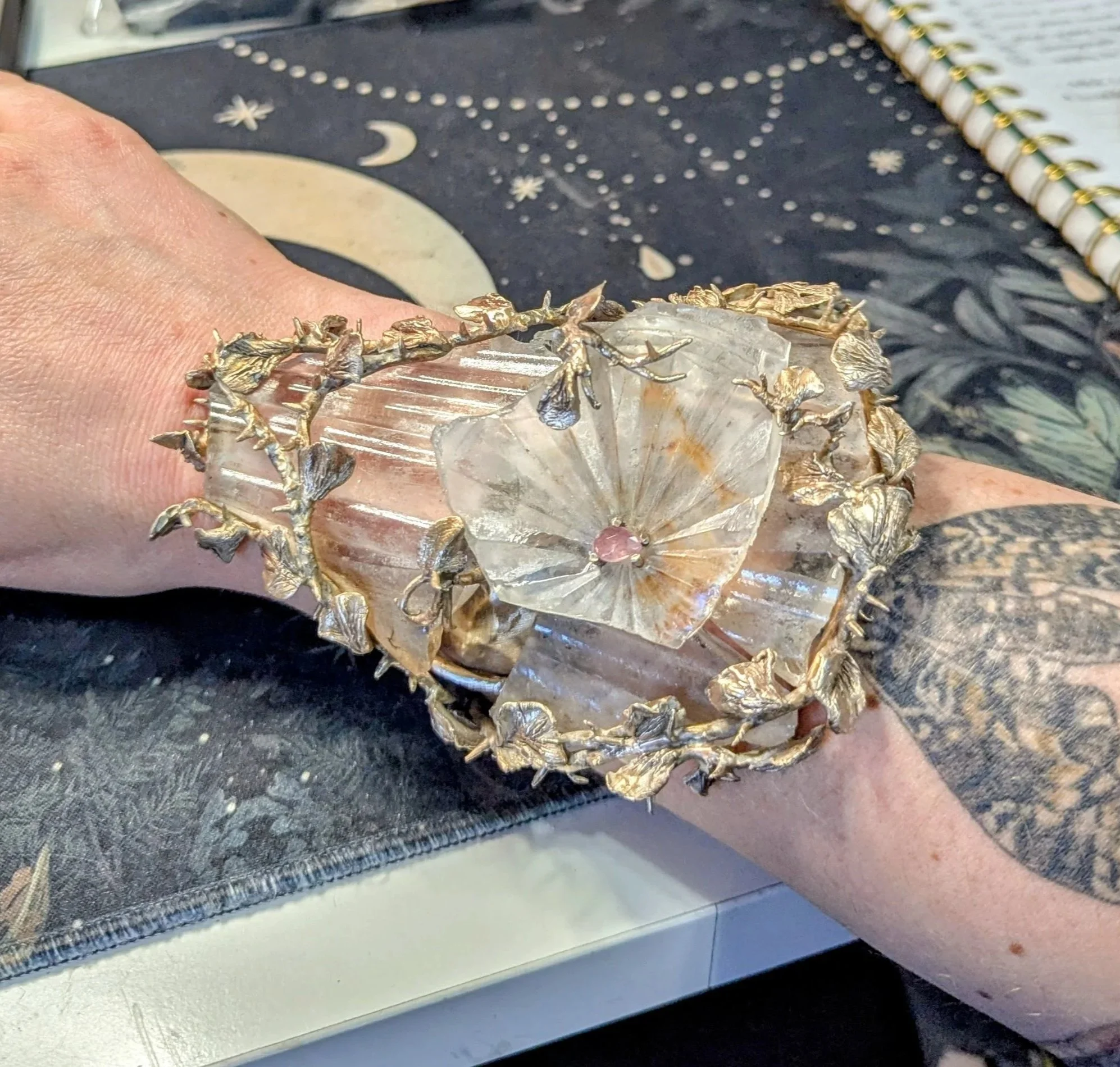 A wrist wearing a cuff bracelet made of bronze, brass and glass with a pink stone set on top