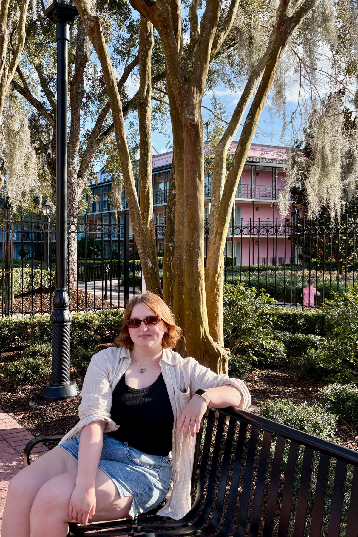 A woman with red hair, sunglasses, and a black shirt sitting on a black park bench in a park, with a tree and buildings in the background.