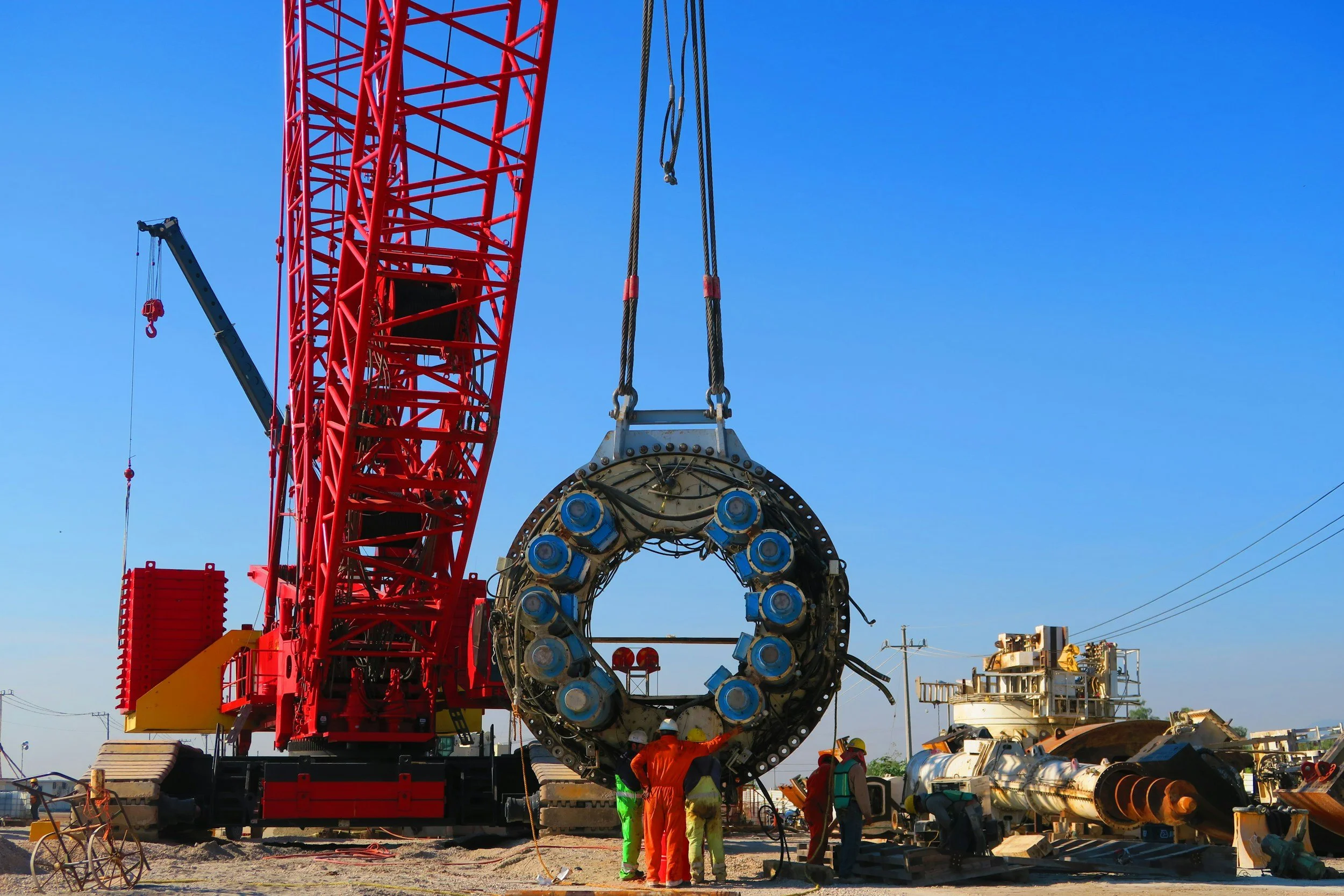 Construction workers in orange and yellow safety gear working on a large drilling machine at a construction site against clear blue sky.