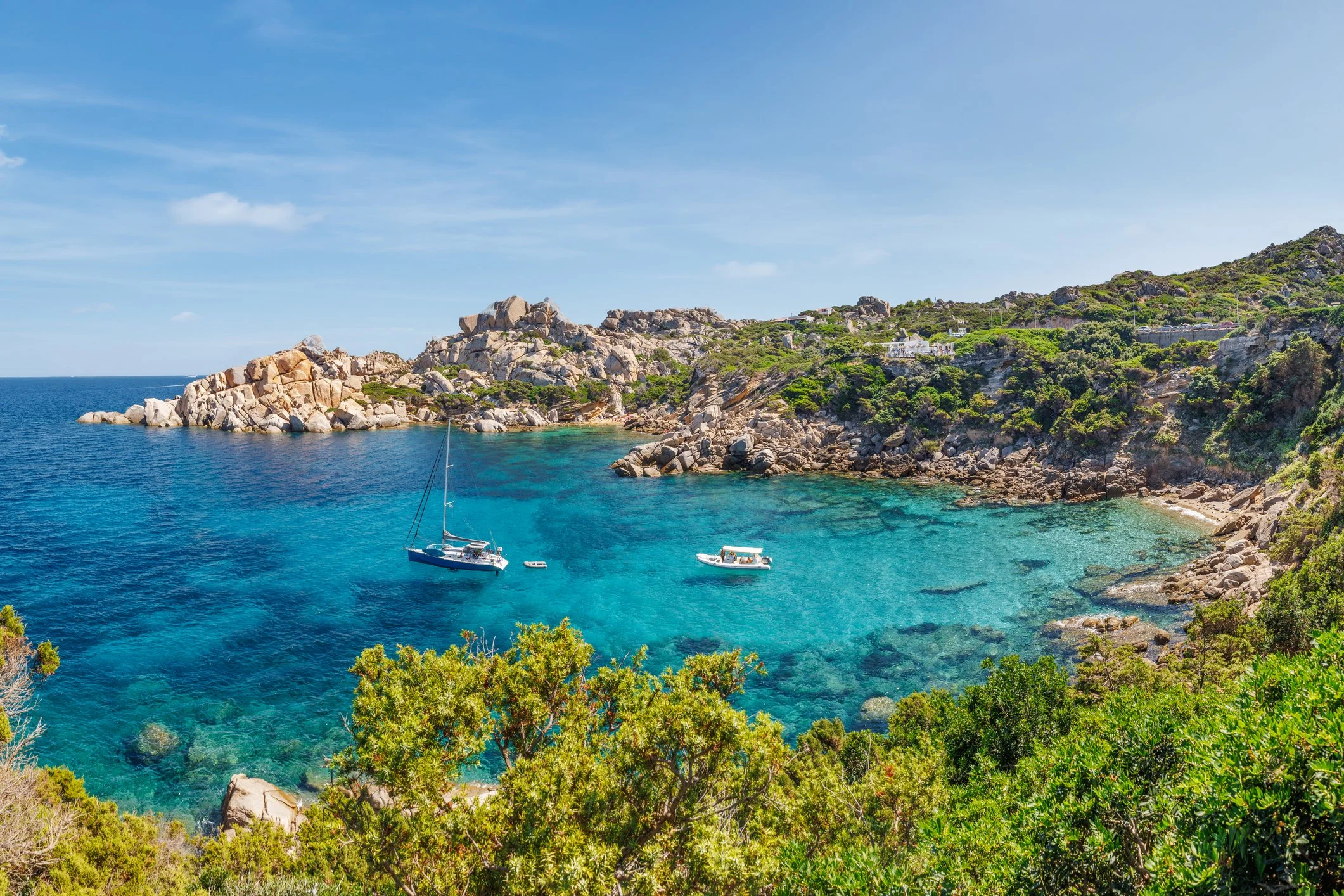 A clear, turquoise bay with two boats anchored near the rocky shore, surrounded by green vegetation and hillside houses under a blue sky.